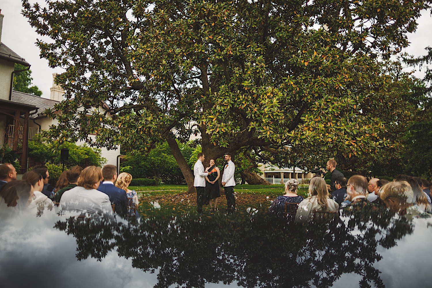 A wedding ceremony is taking place outdoors under a large tree, with a few guests seated and facing the couple, who are flanked by the officiant. The sky is reflected at the bottom of the image, creating an ethereal atmosphere that feels almost like an auto draft for a perfect moment.