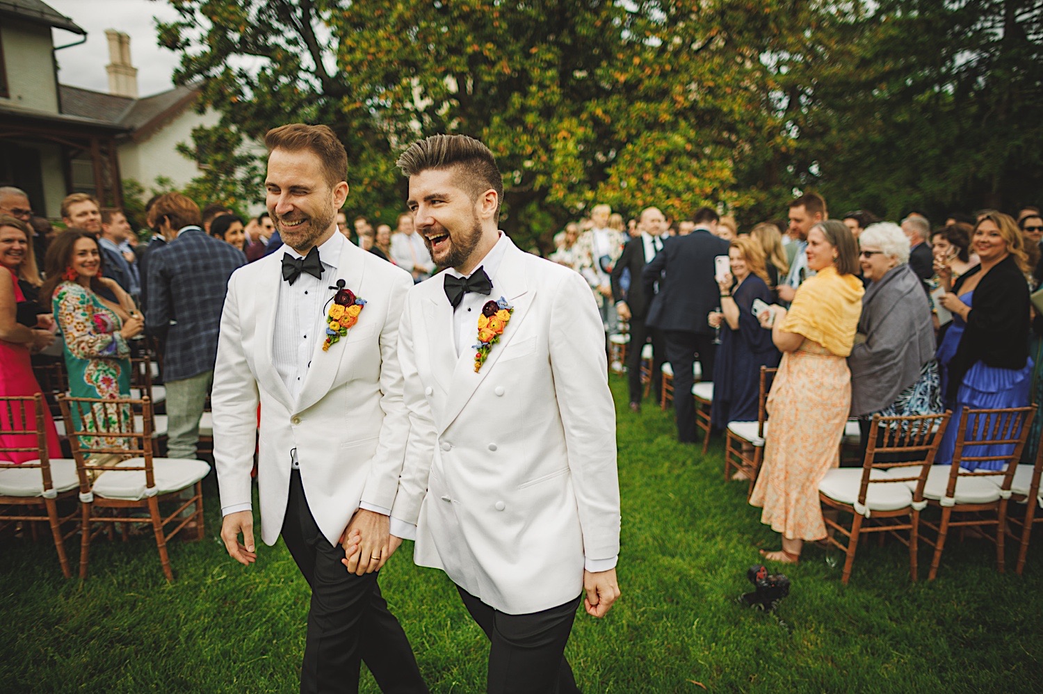 Two men in matching white tuxedos walk hand-in-hand down an outdoor aisle, surrounded by smiling guests and the simplicity of an auto draft guiding their steps.