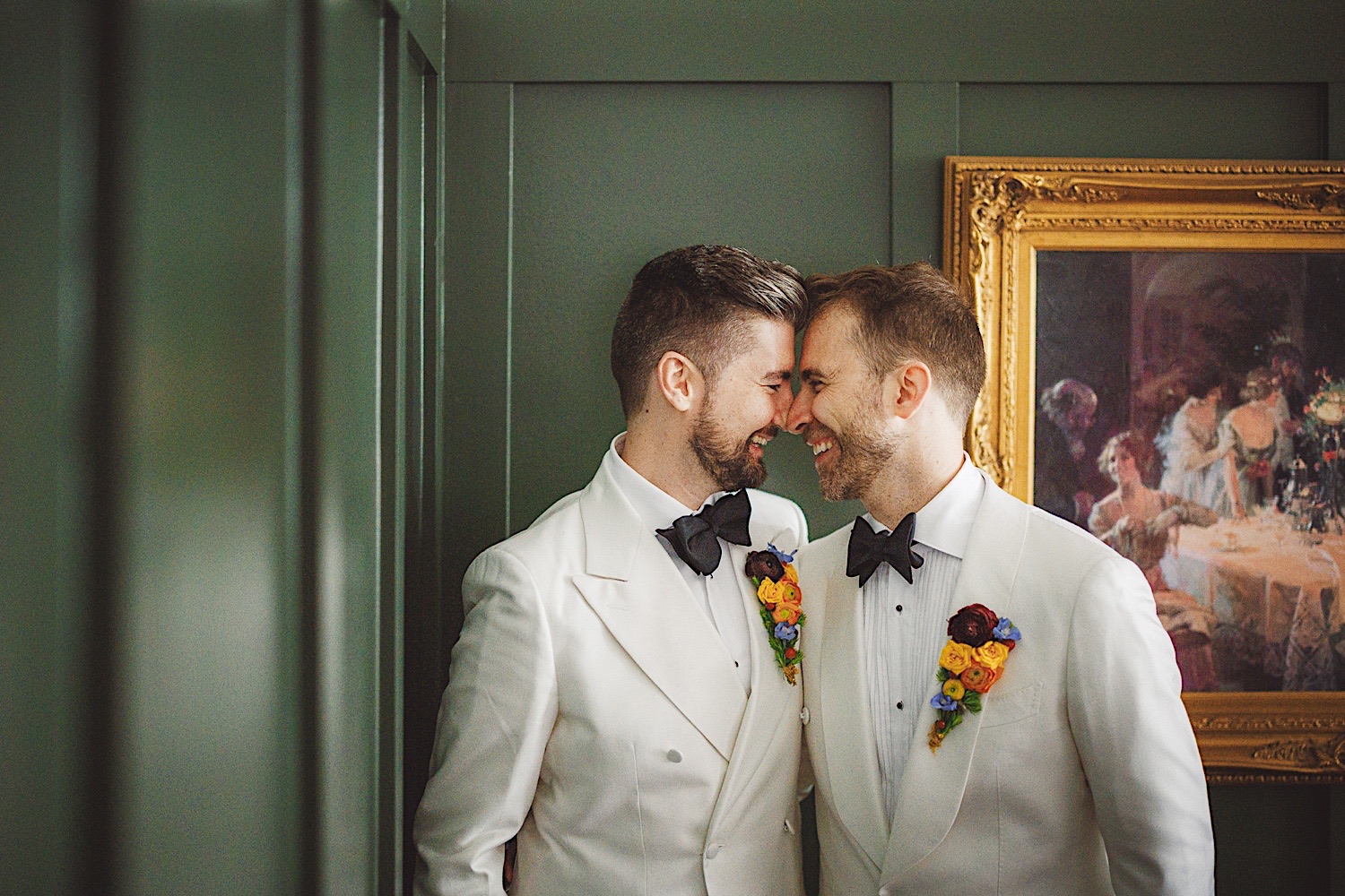 Two men in white tuxedos, adorned with colorful boutonnières, stand closely together, touching foreheads and smiling. They pose in front of a green wall and a framed painting, capturing this moment like an auto draft of pure joy.