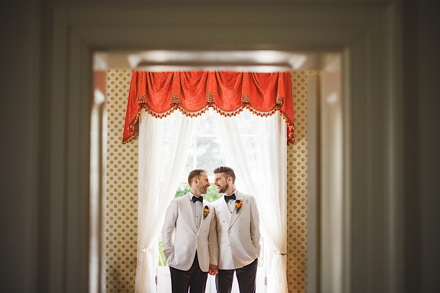 Two men in white tuxedos hold hands and smile at each other in front of a window with red and gold curtains, as if a special moment had been auto-drafted by fate.