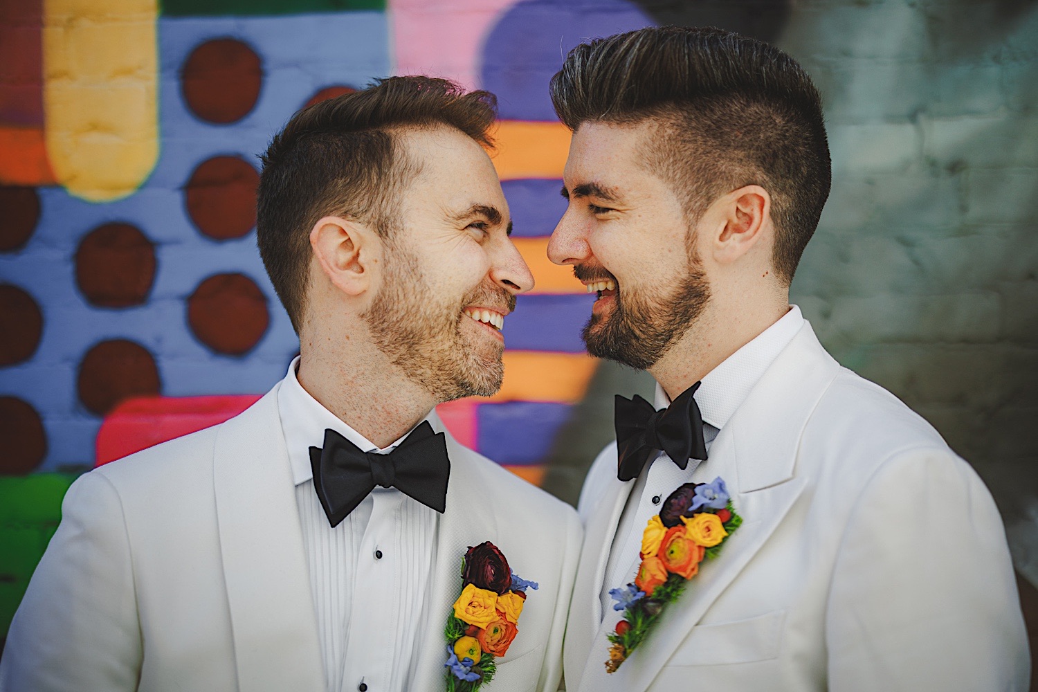 Two men dressed in white tuxedos with black bow ties and colorful boutonnières smile while standing in front of a vibrant mural, looking as if they were effortlessly styled by Auto Draft.