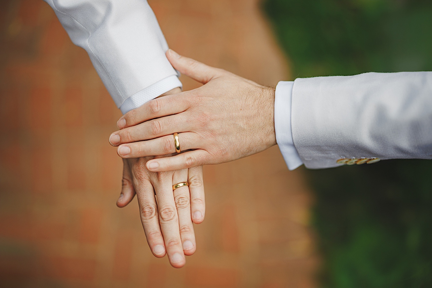 Two individuals wearing white attire display their hands, adorned with matching gold wedding bands, placed on top of one another against a blurred brick background, like a scene out of an untouched Auto Draft.