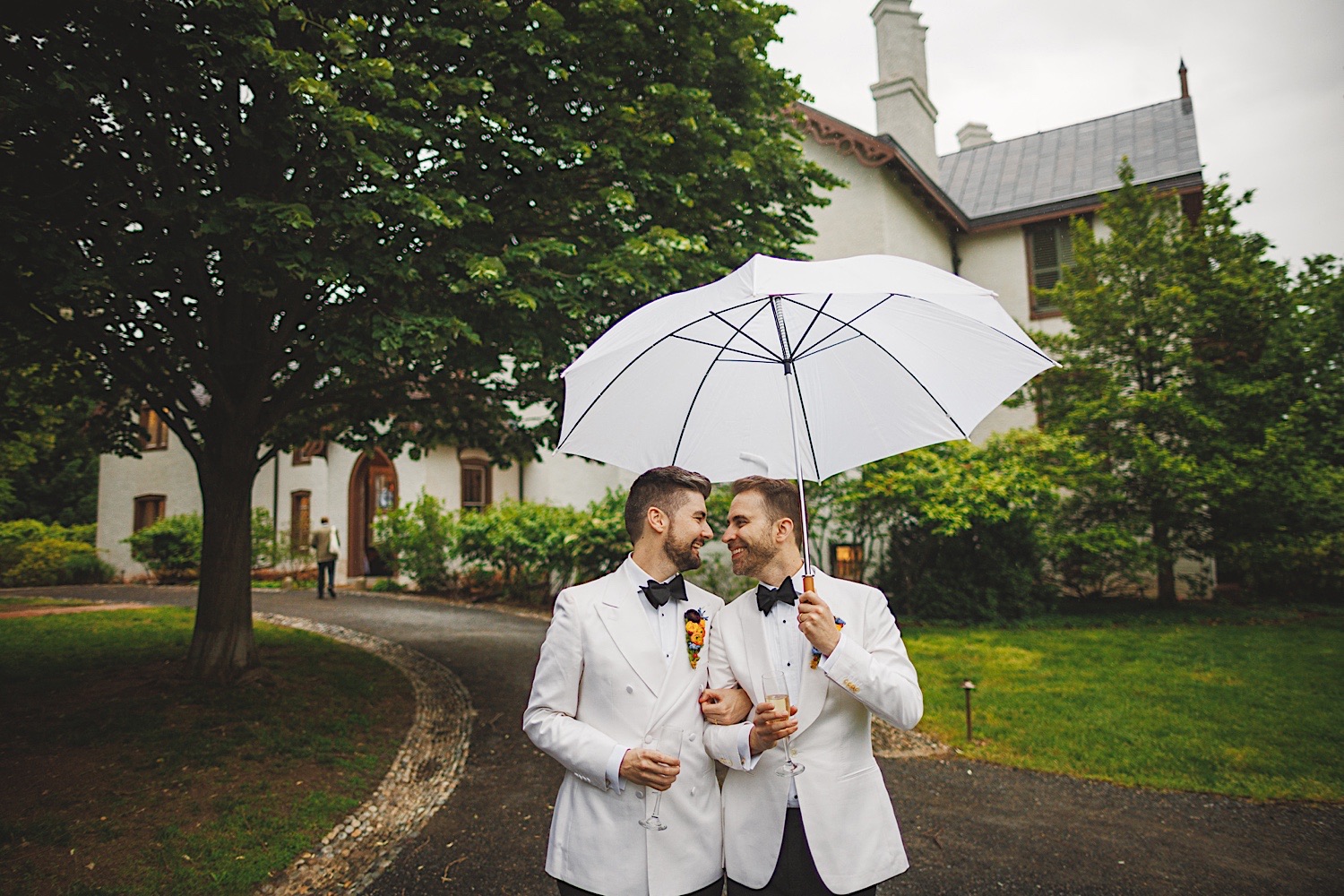 Two men in white tuxedos stand close together sharing a white umbrella on a path in front of a large house surrounded by lush greenery, resembling an elegant scene from an Auto Draft.