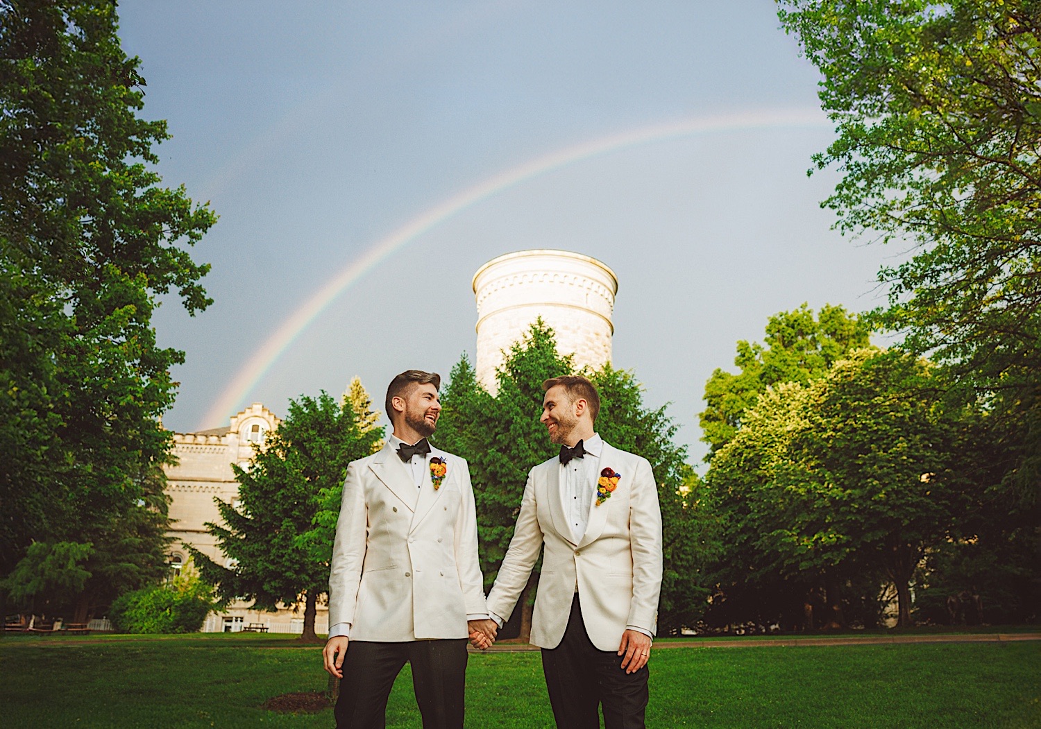 Two men in white suits hold hands and smile at each other in a green park with a double rainbow and a tower in the background, almost as if nature itself approved this perfect auto draft of love.