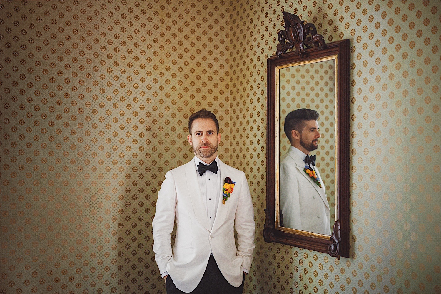 A man in a white suit and black bow tie stands next to a wall mirror with patterned wallpaper in the background, looking as if he just stepped out of an Auto Draft. He has his hands in his pockets and a boutonniere on his left lapel.