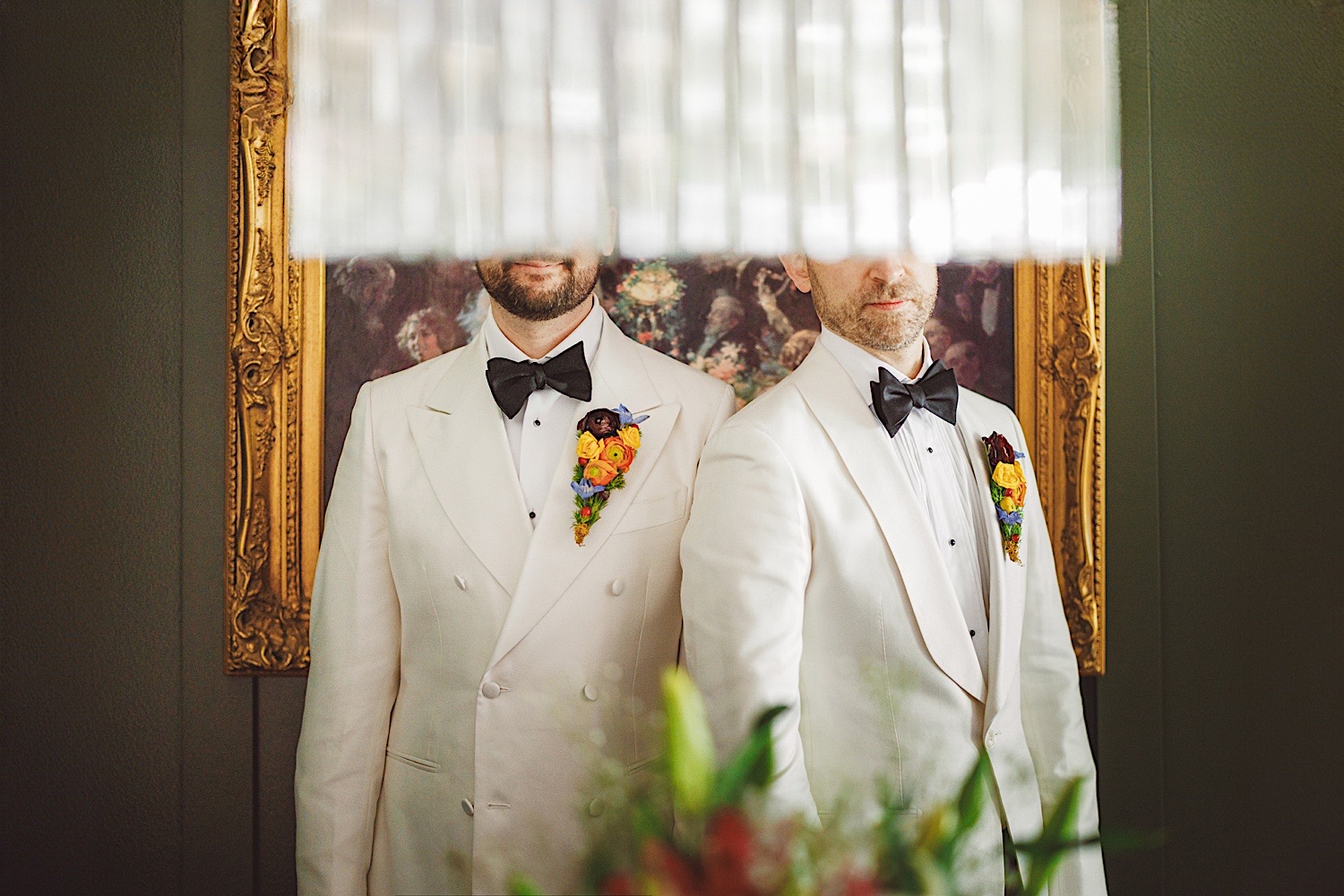 Two men wearing white tuxedos with colorful floral boutonnieres stand side by side; their faces are partially obscured by a hanging light fixture, like an auto draft of an intimate moment caught midway.