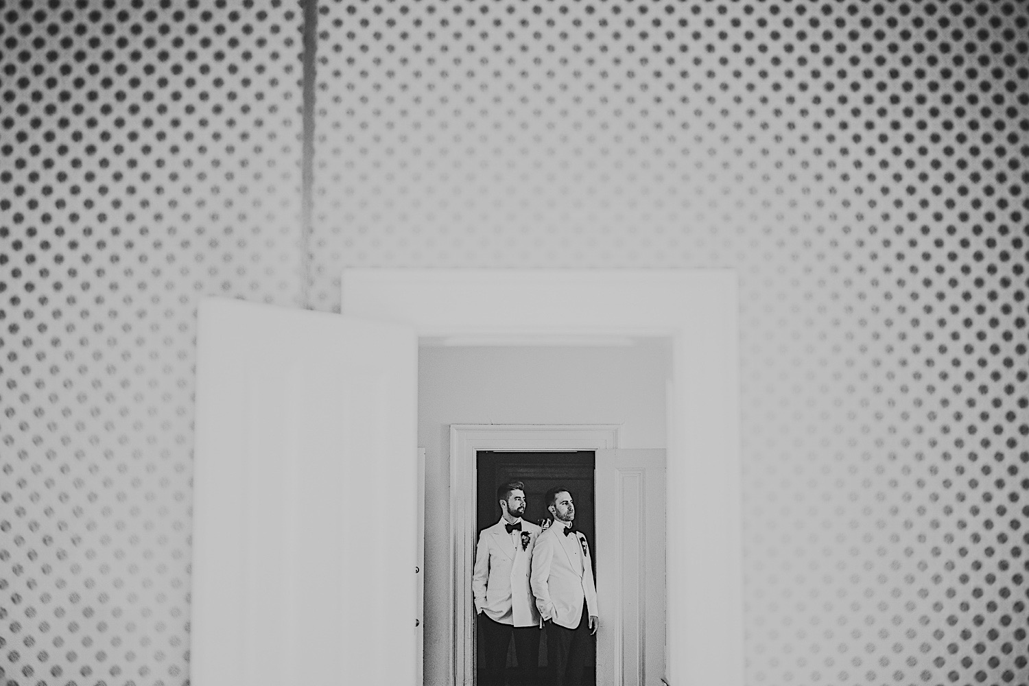 Two men in white tuxedos and bow ties stand side by side, reflected in a doorway mirror. The room features a polka dot wall pattern, almost as if it had been an auto draft of whimsical design ideas.