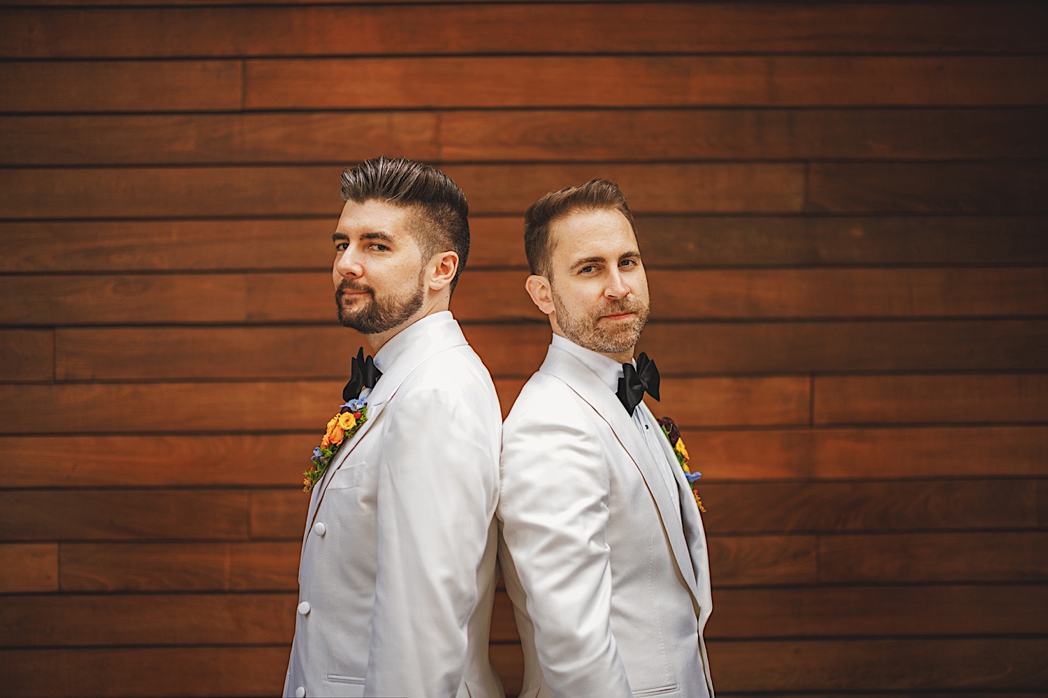 Two men in white tuxedos stand back-to-back in front of a wooden wall, each wearing a black bow tie and a floral boutonniere. It's as if they were meticulously chosen from an Auto Draft of distinguished gentlemen.