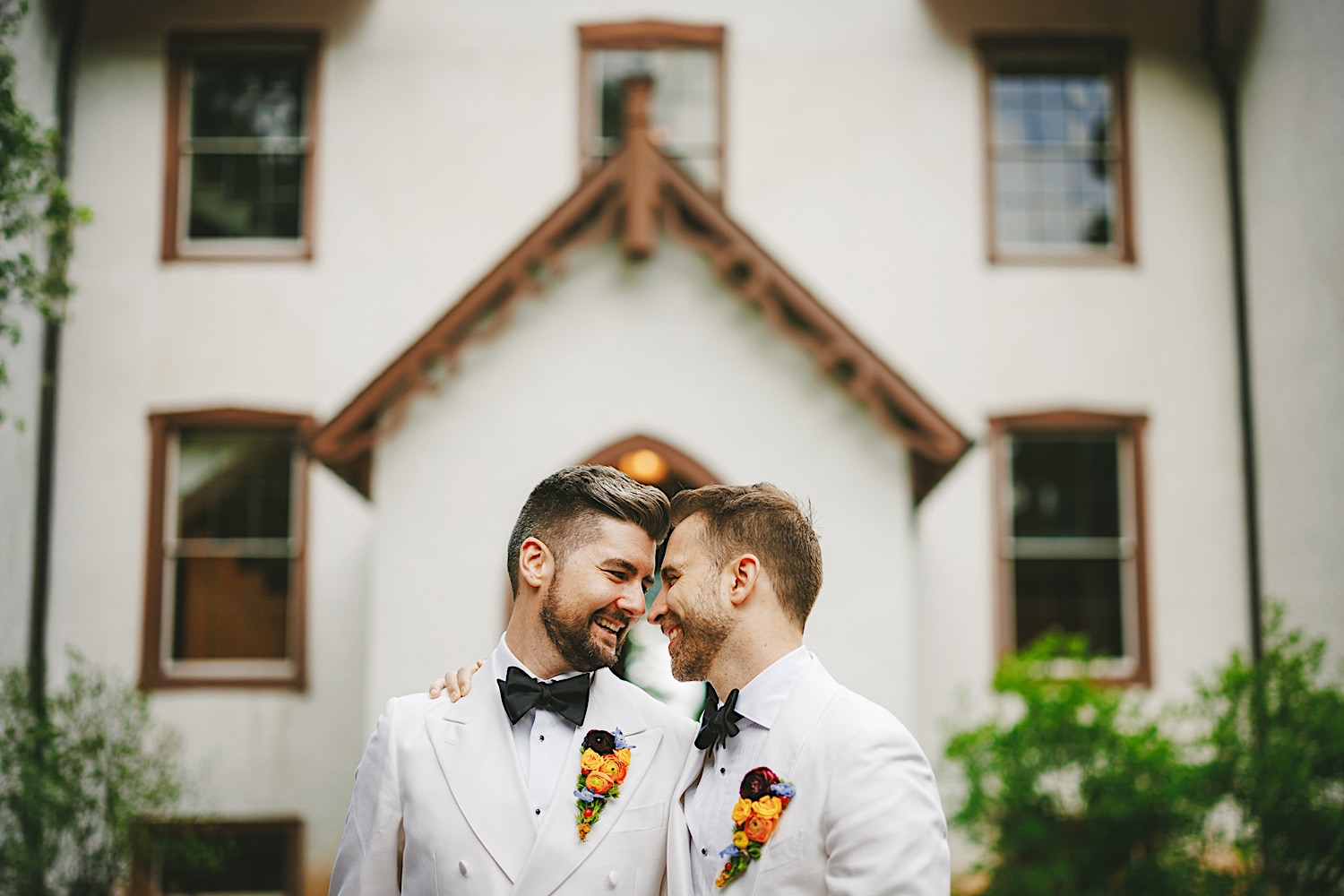 Two men wearing white tuxedos with colorful boutonnieres share a close moment in front of a white house with brown trim, capturing a precious memory that feels almost like an auto draft ready to be polished into a perfect story.