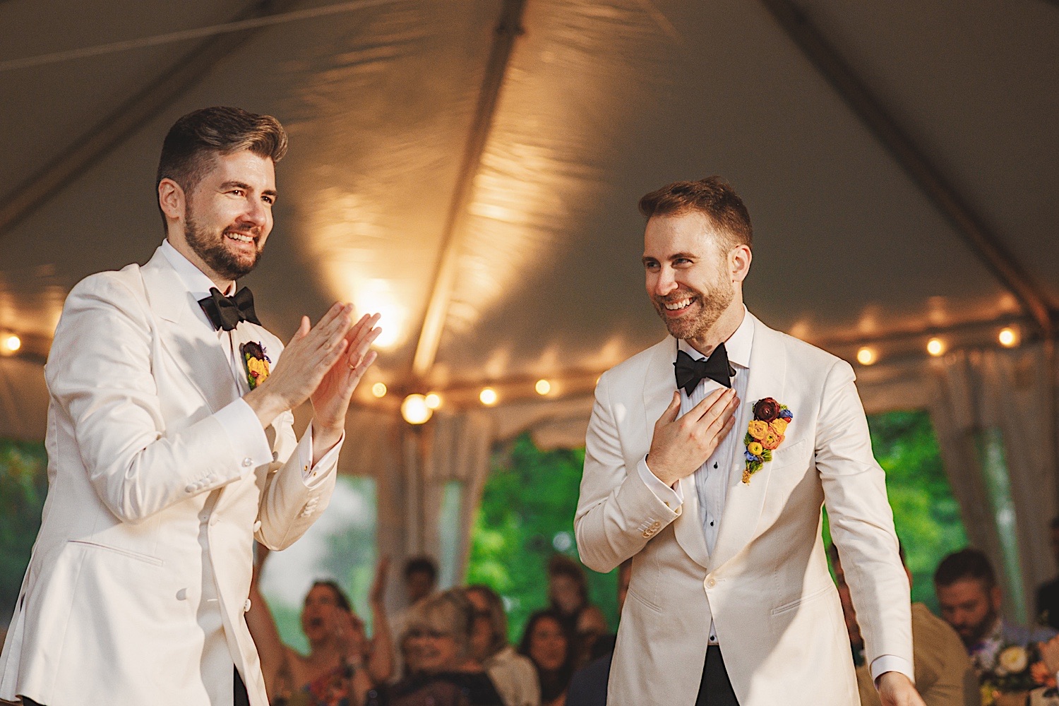 Two grooms in white tuxedos with dark bow ties smiling and applauding at a wedding reception under a white tent, with seated guests in the background. The joyous scene could easily be mistaken for an auto draft from a storybook romance.