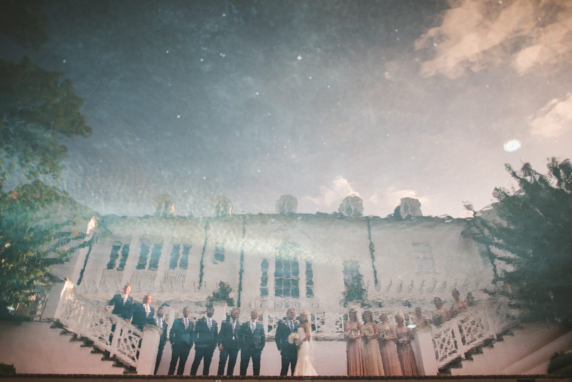 Reflection of a wedding party in water, capturing the essence of wedding photography with a white building with columns in the background, blue sky, and clouds. The group includes the bride, groom, bridesmaids, and groomsmen standing in a line. Contact us for our pricing guide.