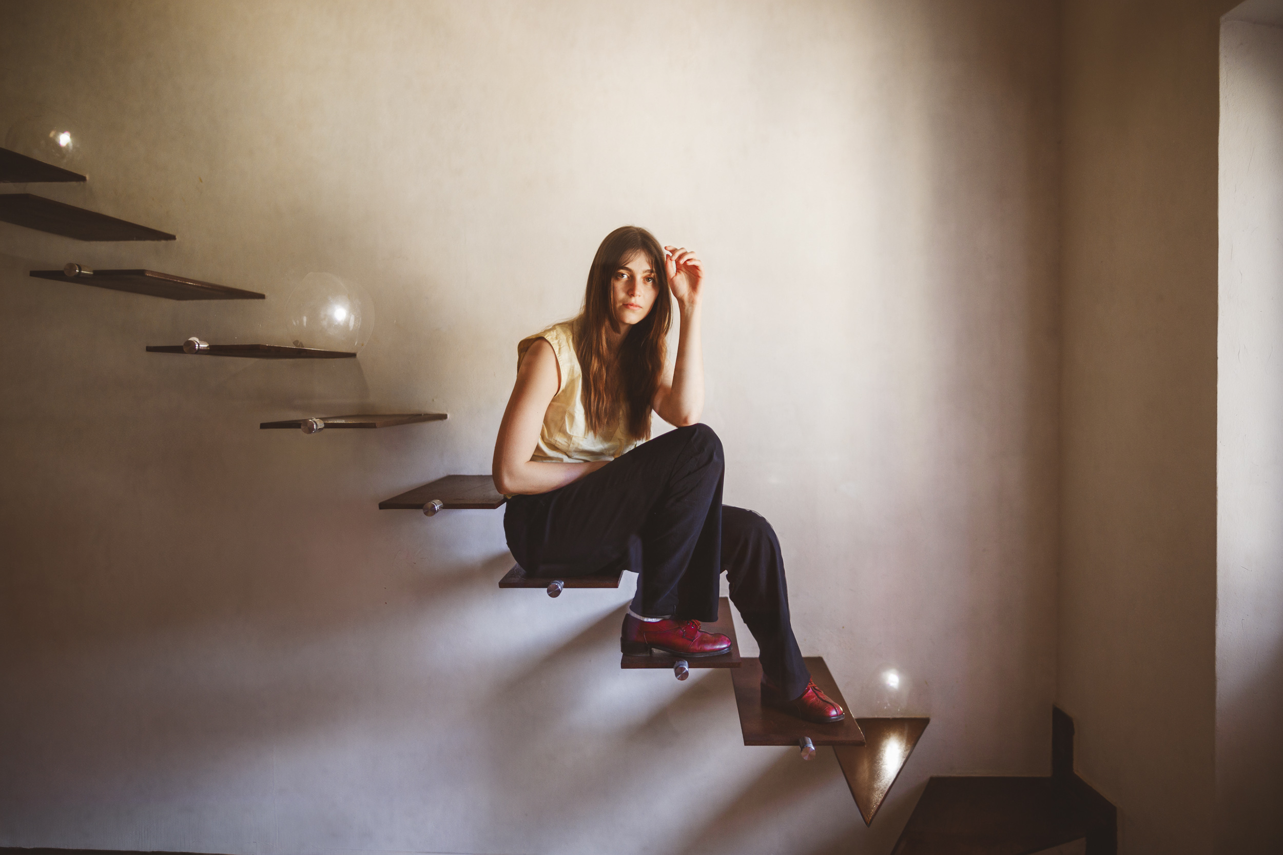 A person sits on a floating staircase in a dimly lit room, wearing dark pants, a light top, and red shoes.