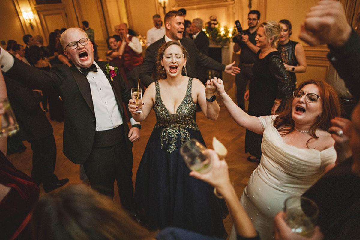 People in formal attire energetically singing and holding drinks at a lively Auto Draft event inside an ornate venue.