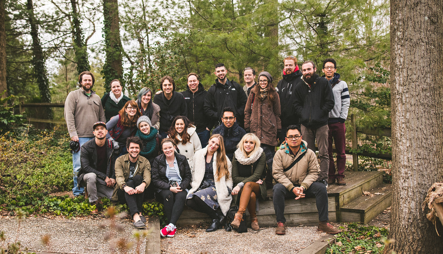 A group of 21 people standing and sitting outdoors in a wooded area, posing for a photo on wooden steps and a gravel path.