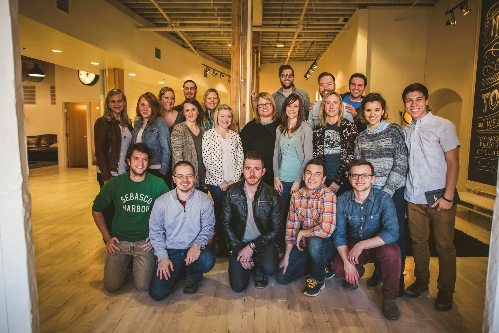 A group of 21 people are standing and kneeling indoors, smiling at the camera. They are casually dressed and arranged in three rows in what appears to be an office space.