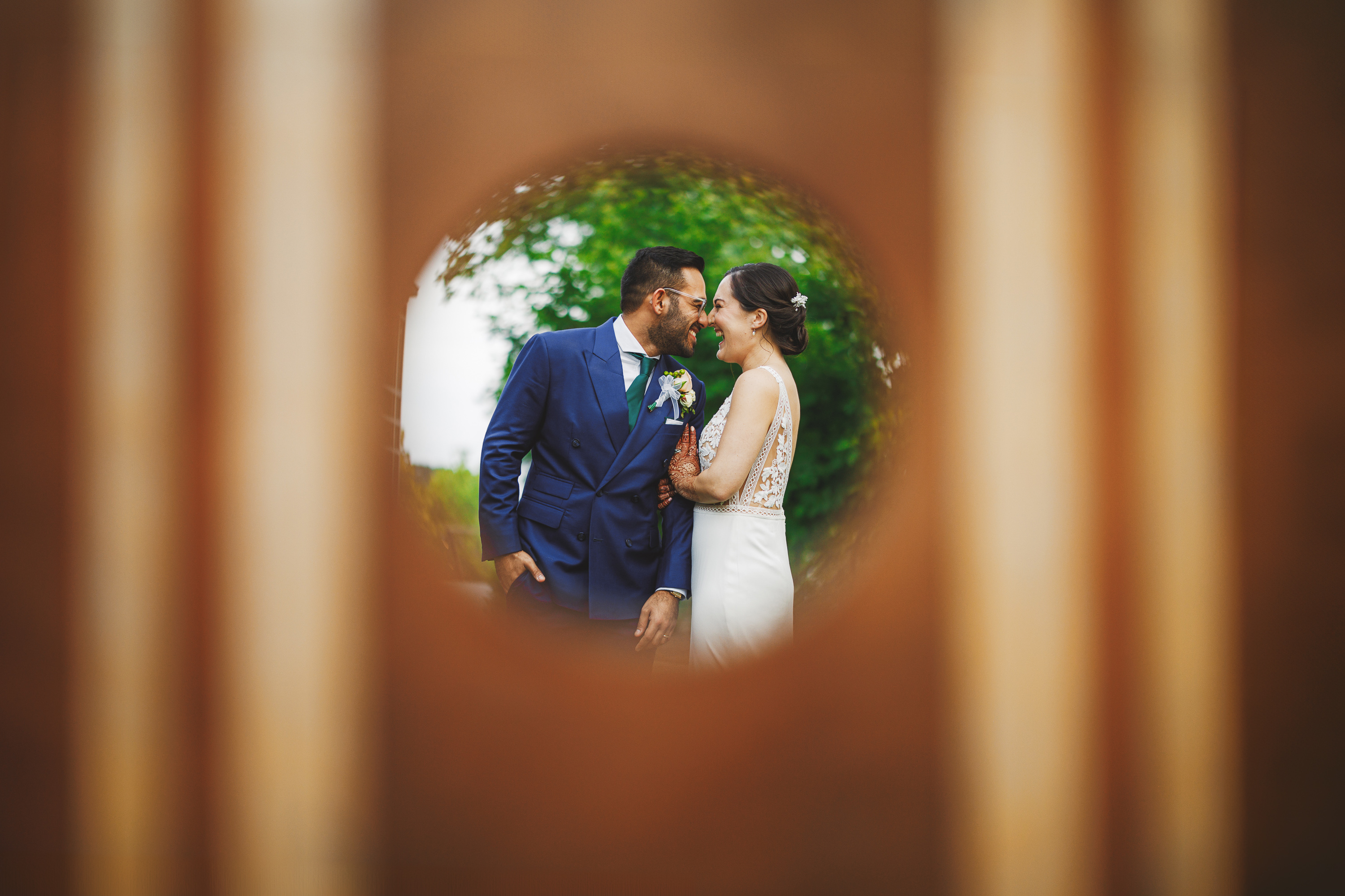 A couple dressed in formal attire stands close together, smiles through a circular wooden frame with a grassy background, captured effortlessly like an auto draft.