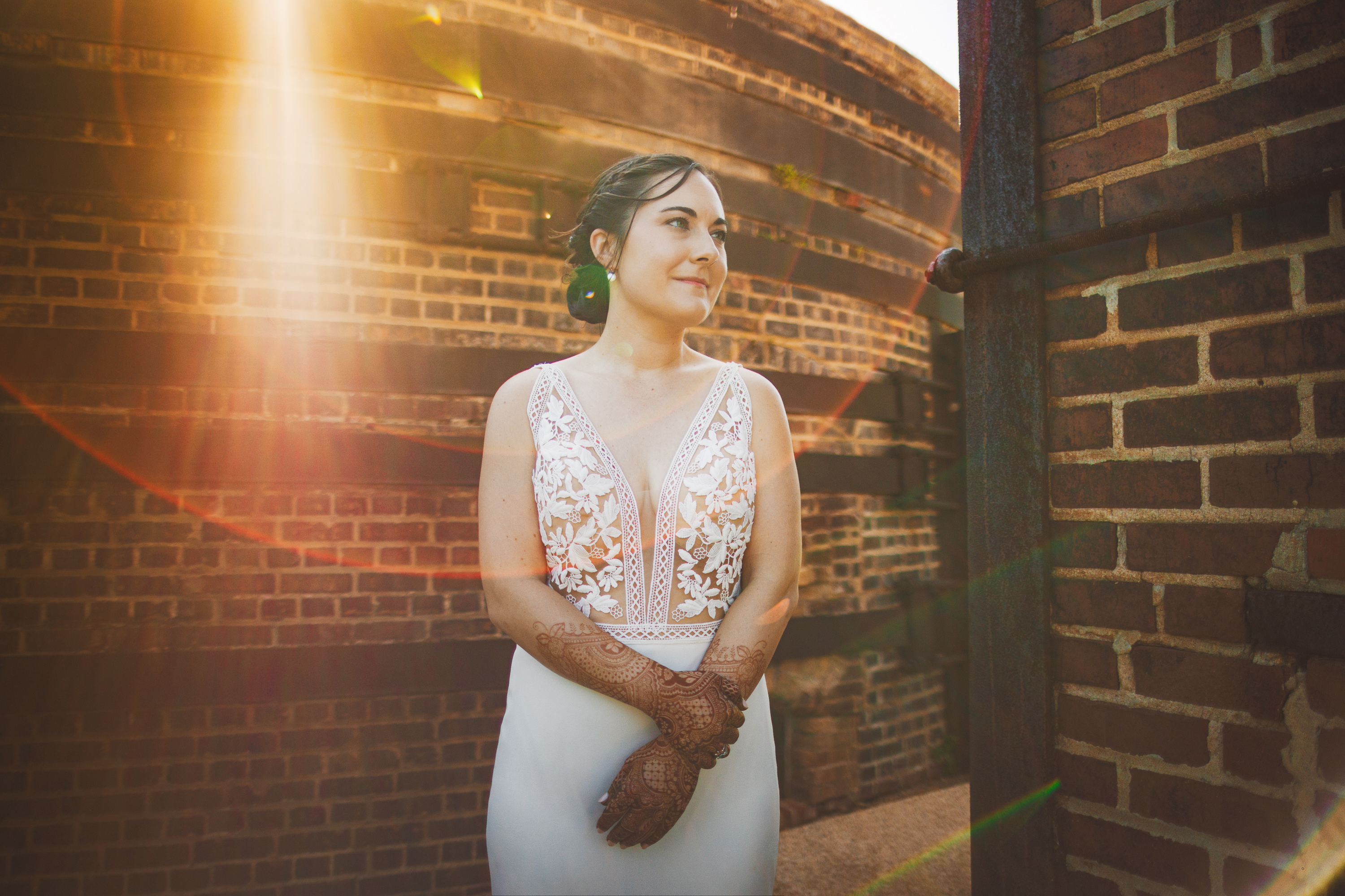 A woman in a white dress with lace details and henna on her hands stands gracefully in front of a brick structure, bathed in sunlight streaming from the top left of the image, as if captured by an auto draft.