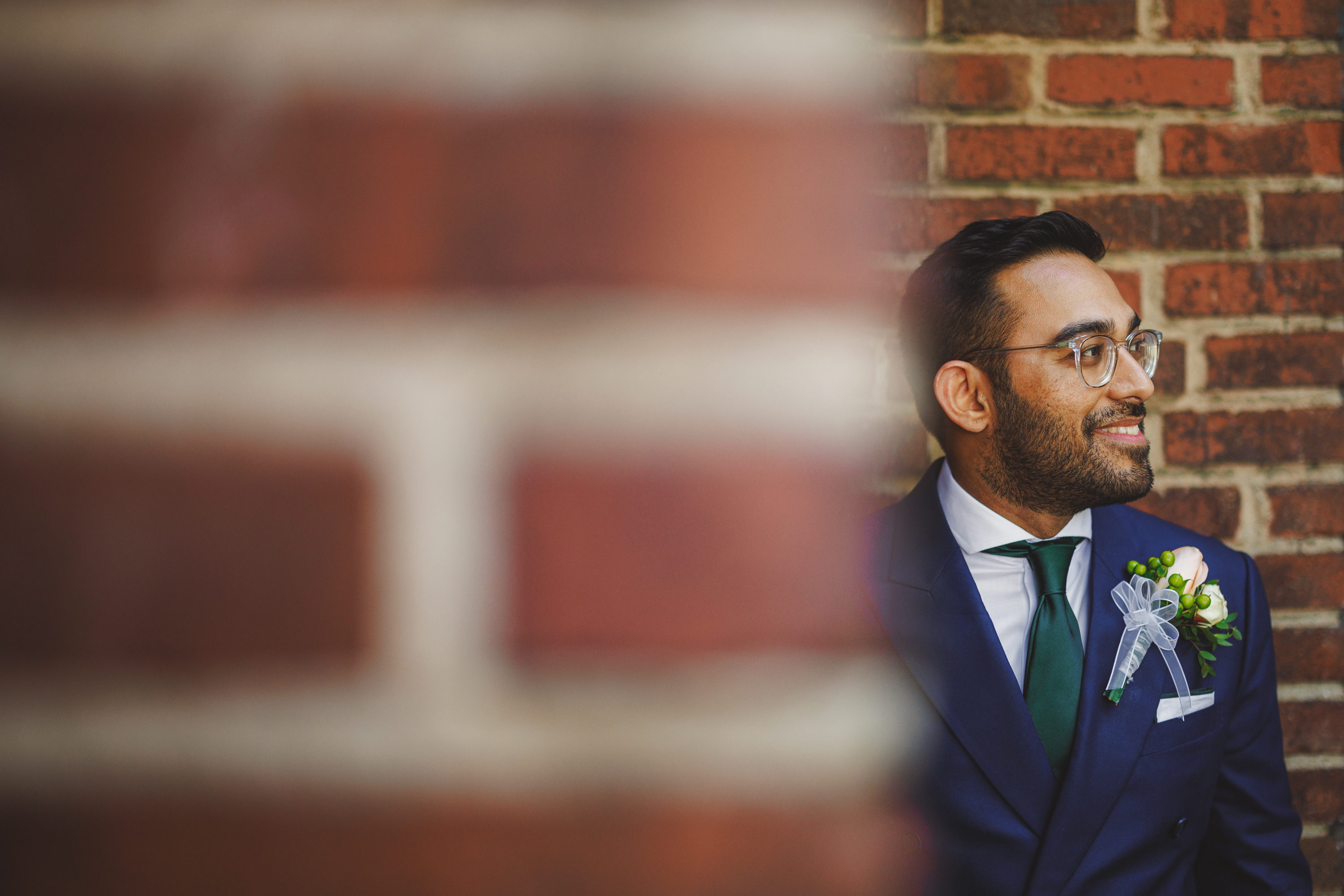 A man in a dark suit with a green tie and boutonnière stands near a brick wall, smiling and looking to the side, as if he's waiting for an auto draft to finalize.