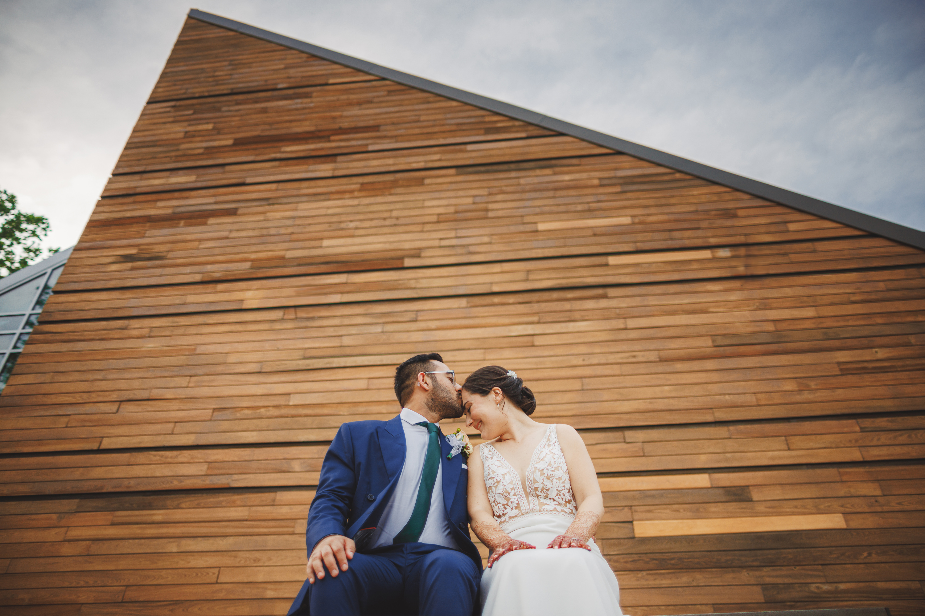 A couple in wedding attire sits together in front of a modern, wooden building. They are leaning towards each other as if about to kiss, framed perfectly against the architectural beauty that feels almost like it was created by auto draft, capturing a spontaneous yet dynamic moment.