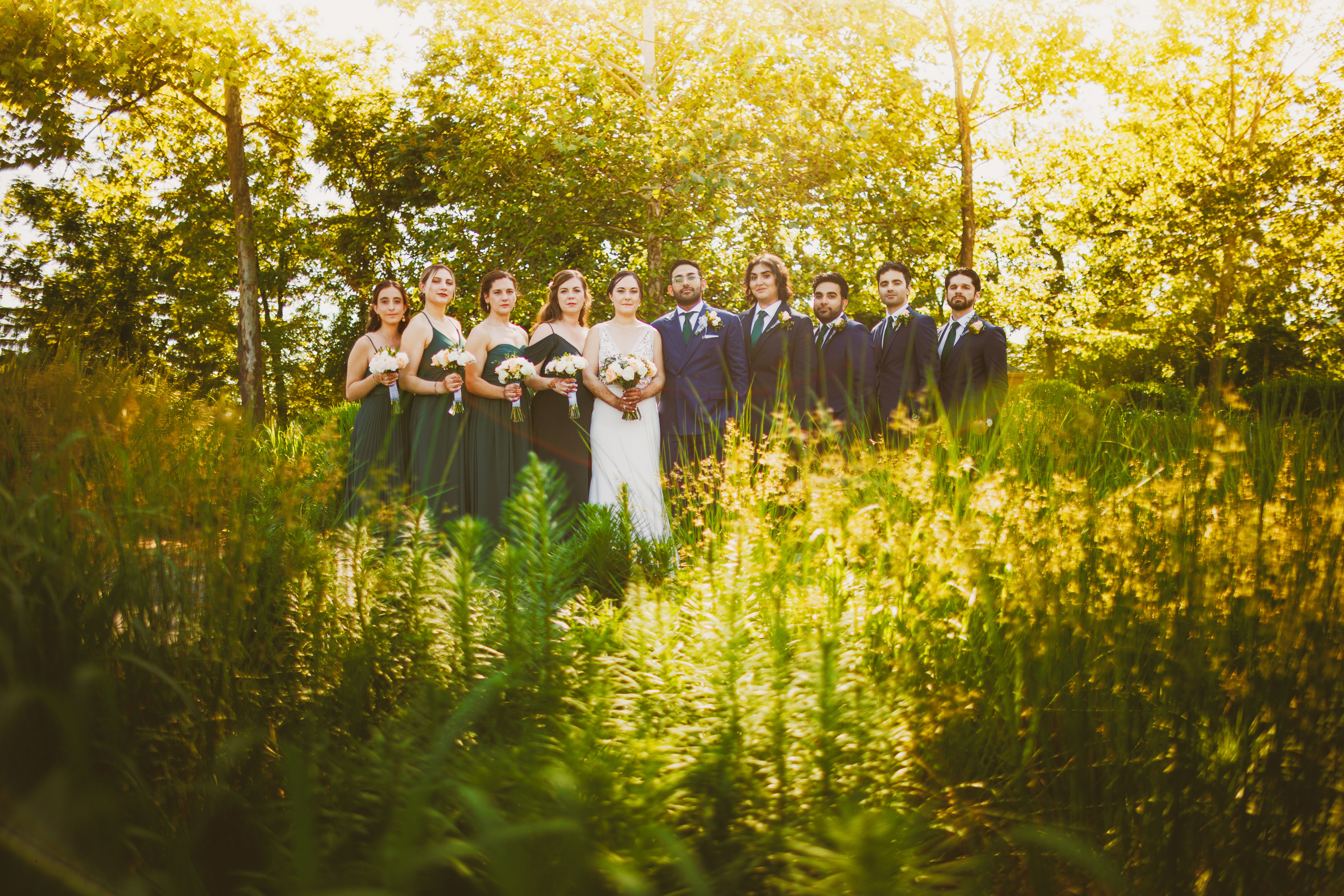A wedding party, consisting of six bridesmaids in green dresses and four groomsmen in dark suits, stands together like an auto draft painting in an outdoor setting surrounded by tall grass and trees under bright sunlight.