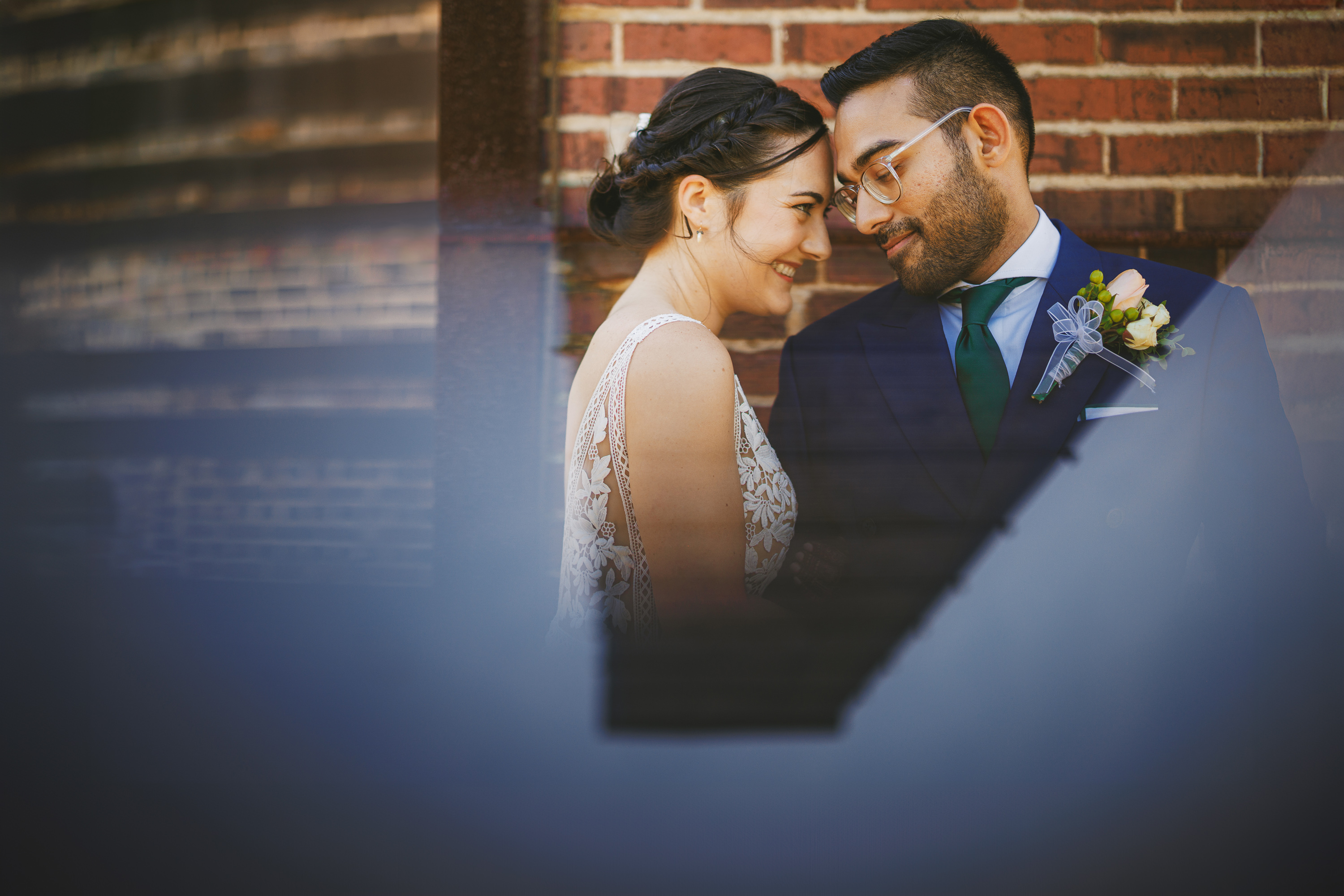 A man and woman in formal attire stand close together outdoors, smiling and looking at each other affectionately against a brick wall background, seemingly captured in an auto draft of love.