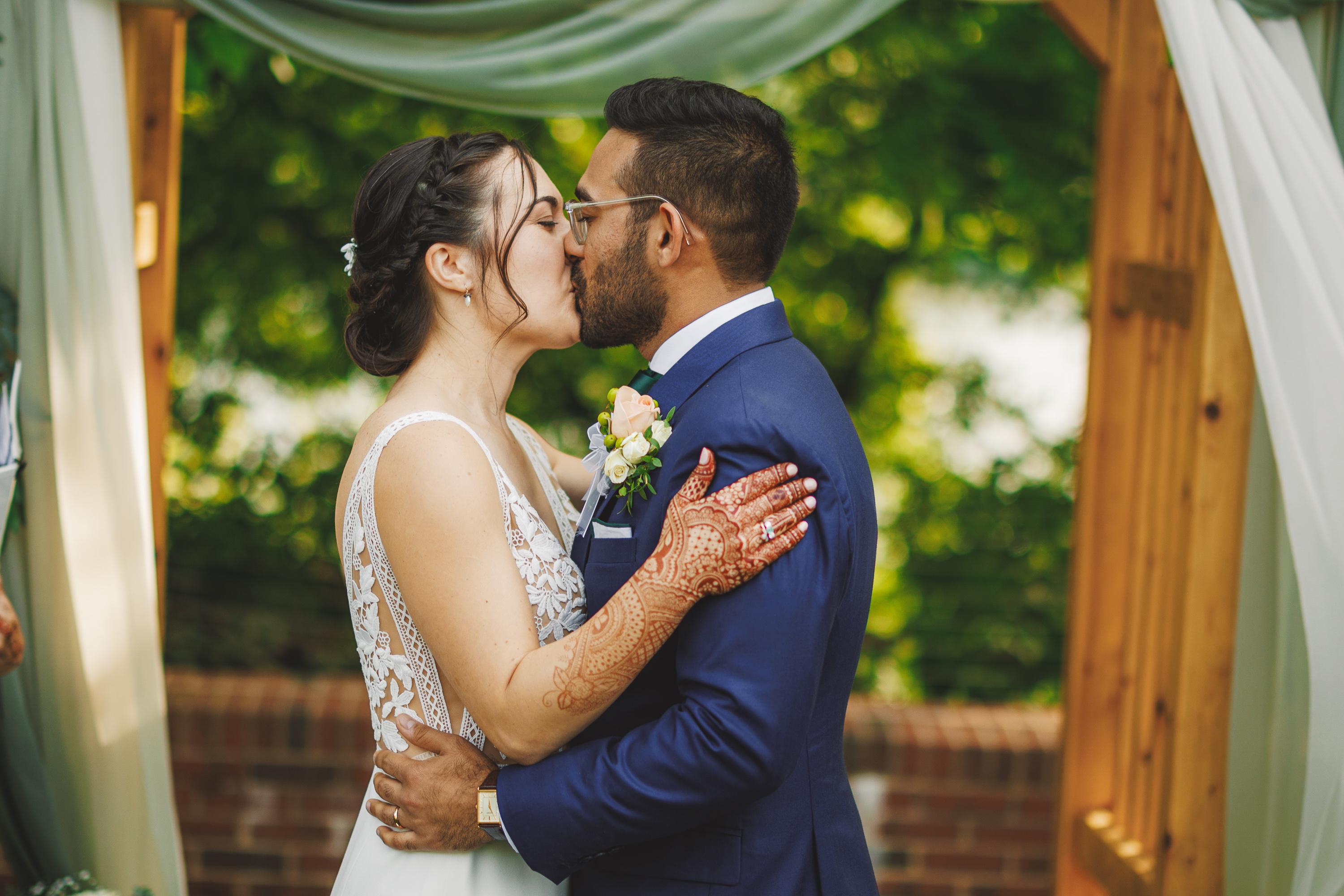A bride in a white gown and a groom in a blue suit share a kiss under an outdoor wooden archway, beautifully draped with white fabric. With henna on her arms, she brings an elegant charm to their special moment.