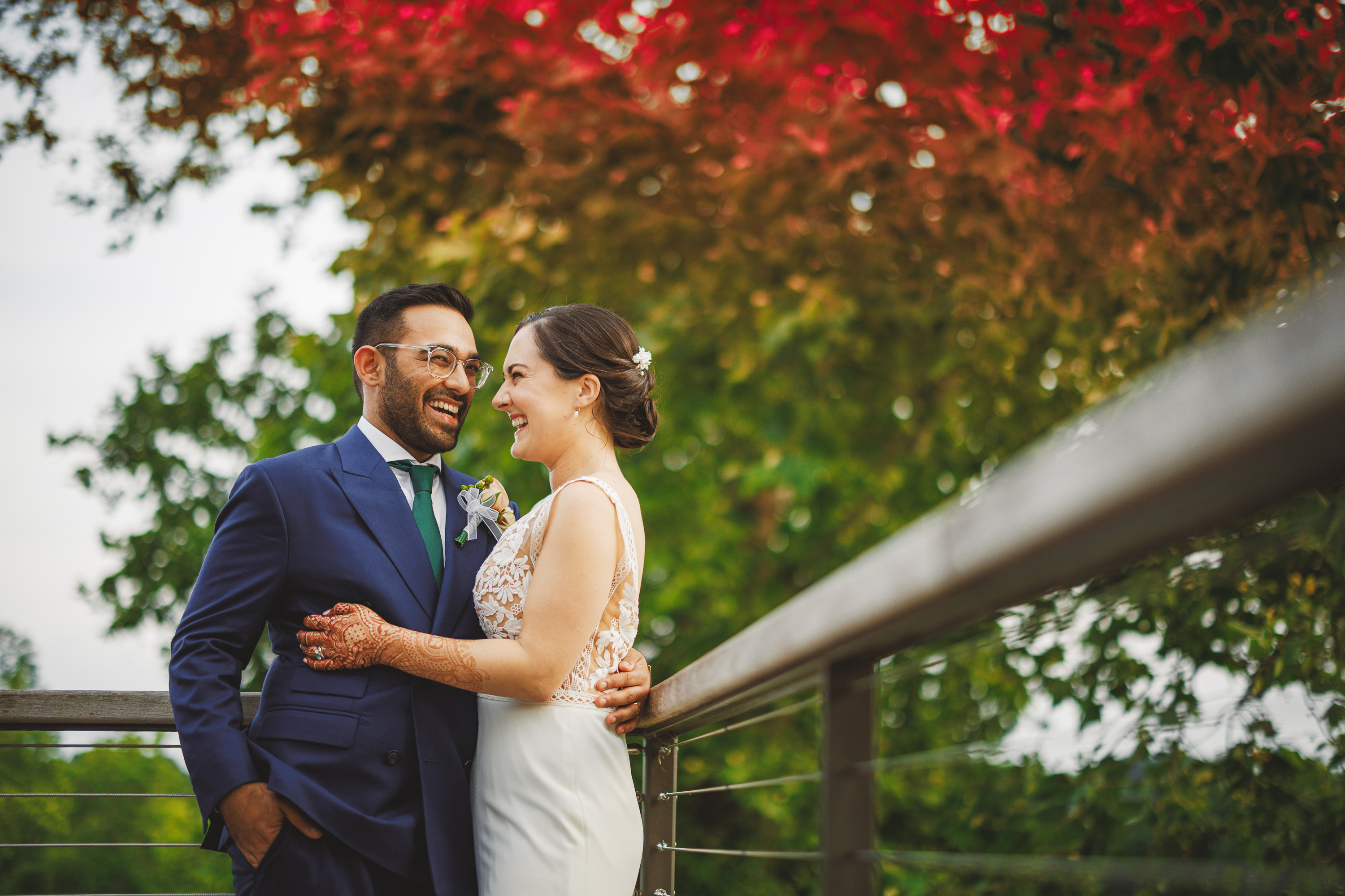 A couple in formal attire embraces on a wooden deck with colorful foliage in the background, as if crafted by Auto Draft. The man wears a navy suit and the woman is in a white dress, both smiling at each other.