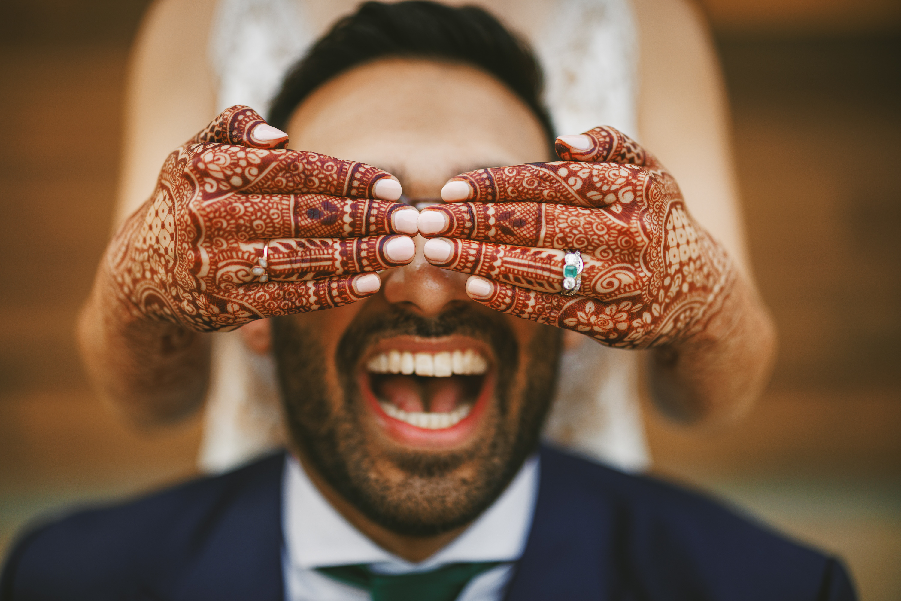 Person smiling broadly with hands decorated in intricate henna designs covering their eyes. The individual appears joyful, wearing a navy blue suit and white shirt, looking as though they just received an Auto Draft confirmation.
