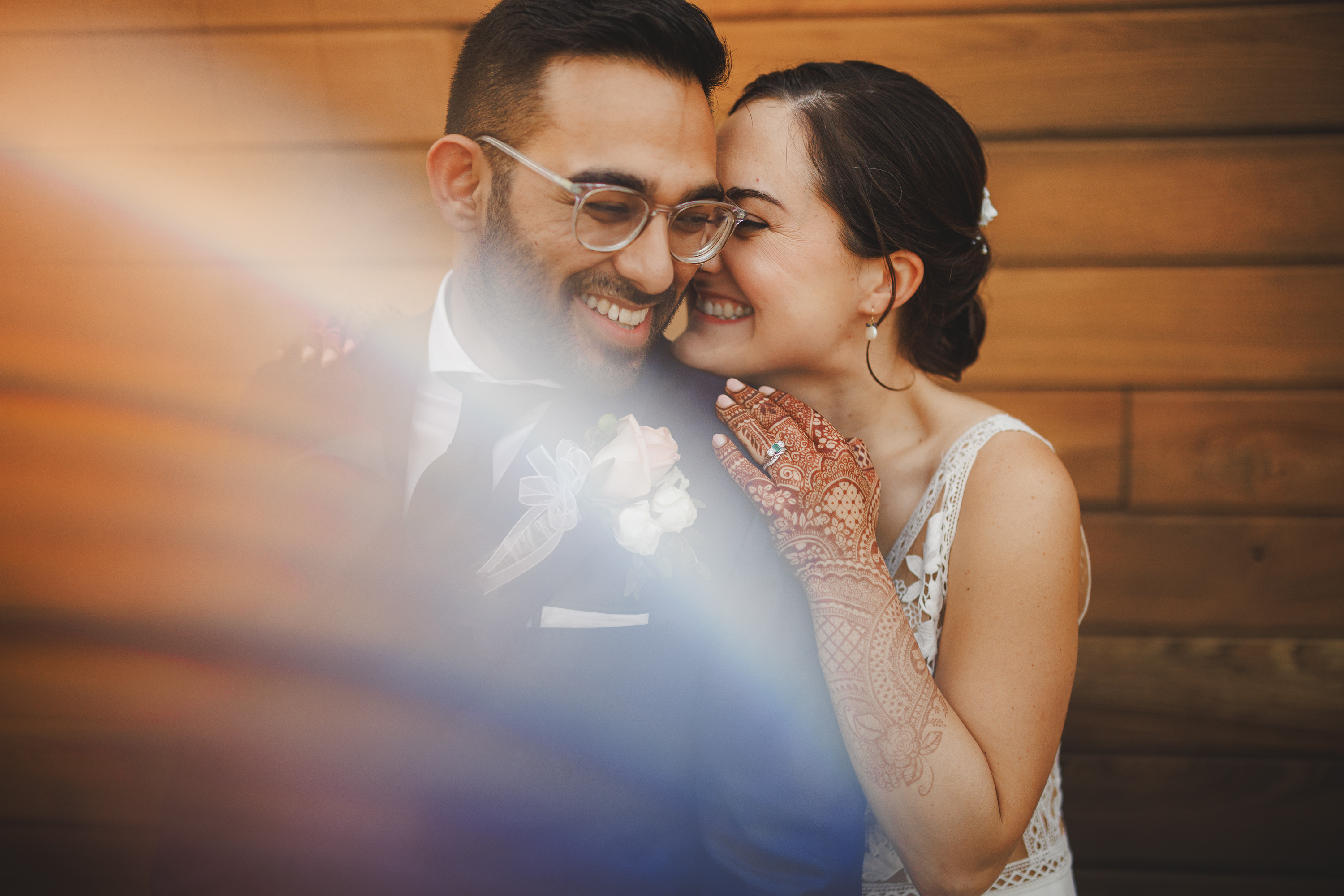 A couple, dressed in wedding attire, smile and embrace each other in front of a wooden backdrop. The bride has henna on her hands. A soft rainbow light effect partially covers the image, giving it an enchanting auto draft quality.