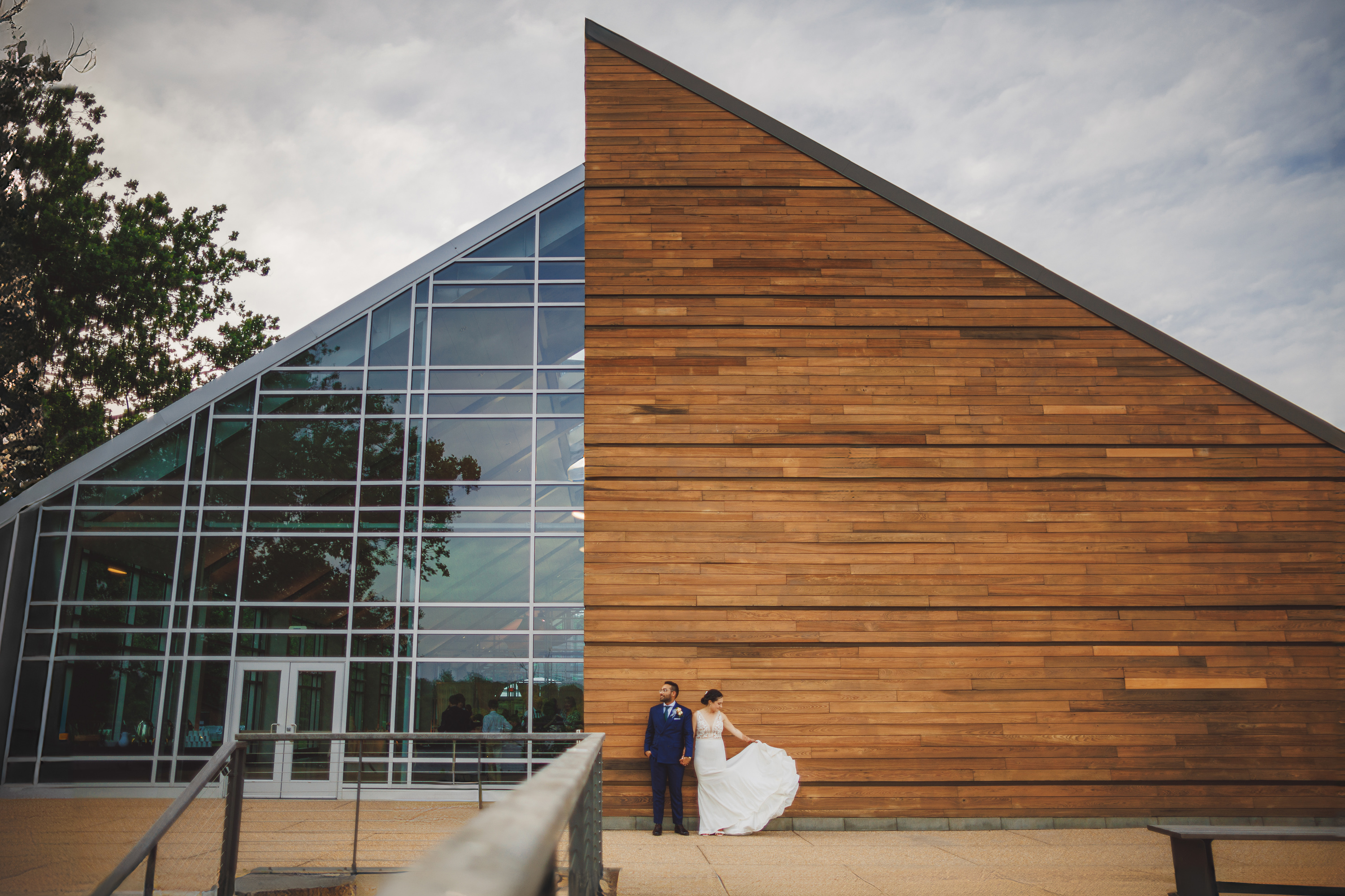 A couple in wedding attire stands in front of a modern building with glass and wooden facades, looking like an Auto Draft from a fairytale. The groom is in a blue suit, and the bride is holding her white gown. Trees add a touch of nature to the left.
