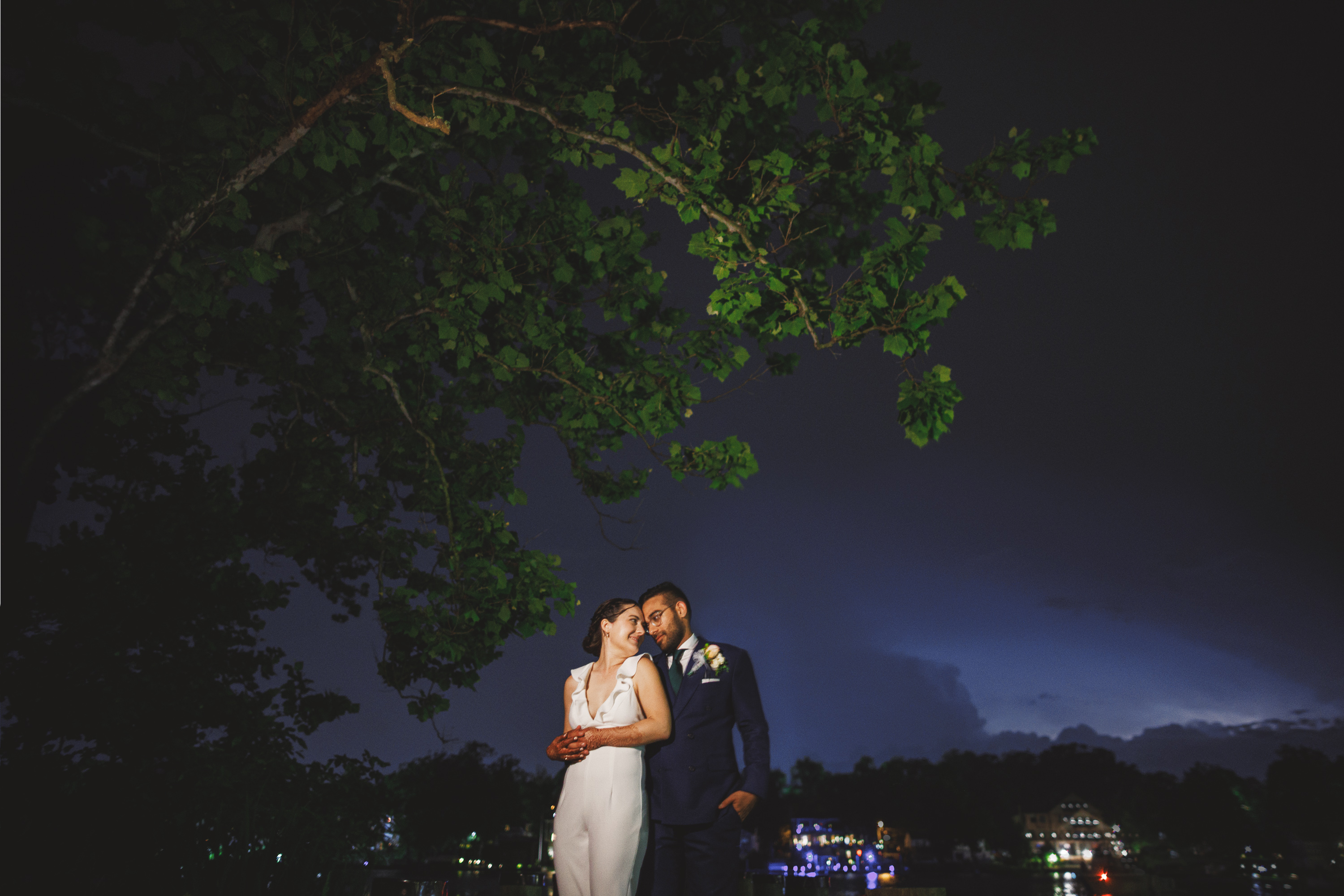 A couple in formal wear stands together outdoors at night under a tree with buildings illuminated in the background, exuding a timeless elegance that feels like an auto draft of a classic romance novel.