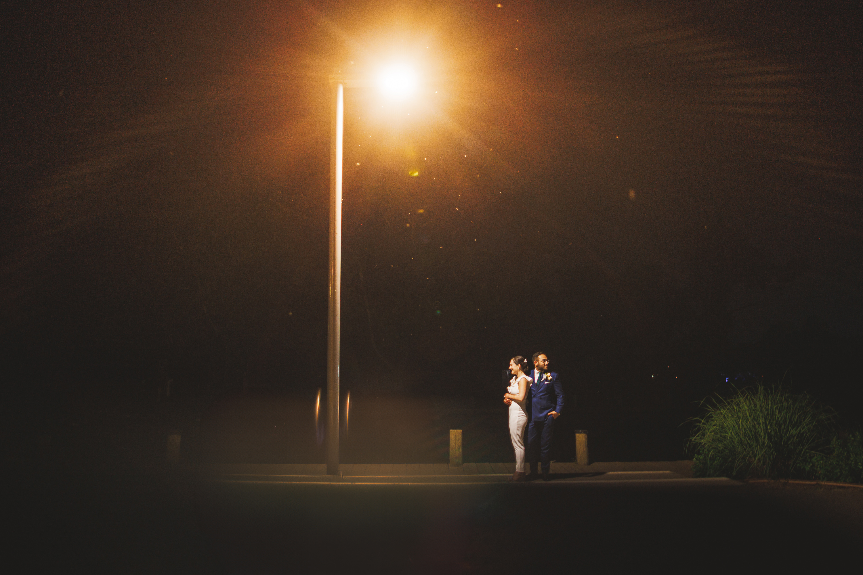 A couple stands close together under a bright streetlight at night, their shadows merging in the dark background, creating a scene reminiscent of an auto draft, with minimal surrounding visibility.
