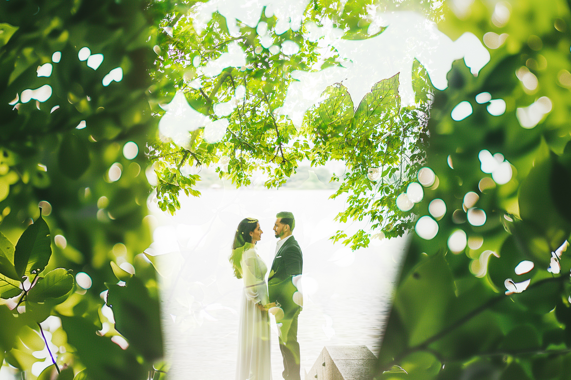 A couple stands facing each other under a canopy of green leaves, with blurred foliage creating a dreamy, almost ethereal effect around them, as if nature itself was on auto draft.