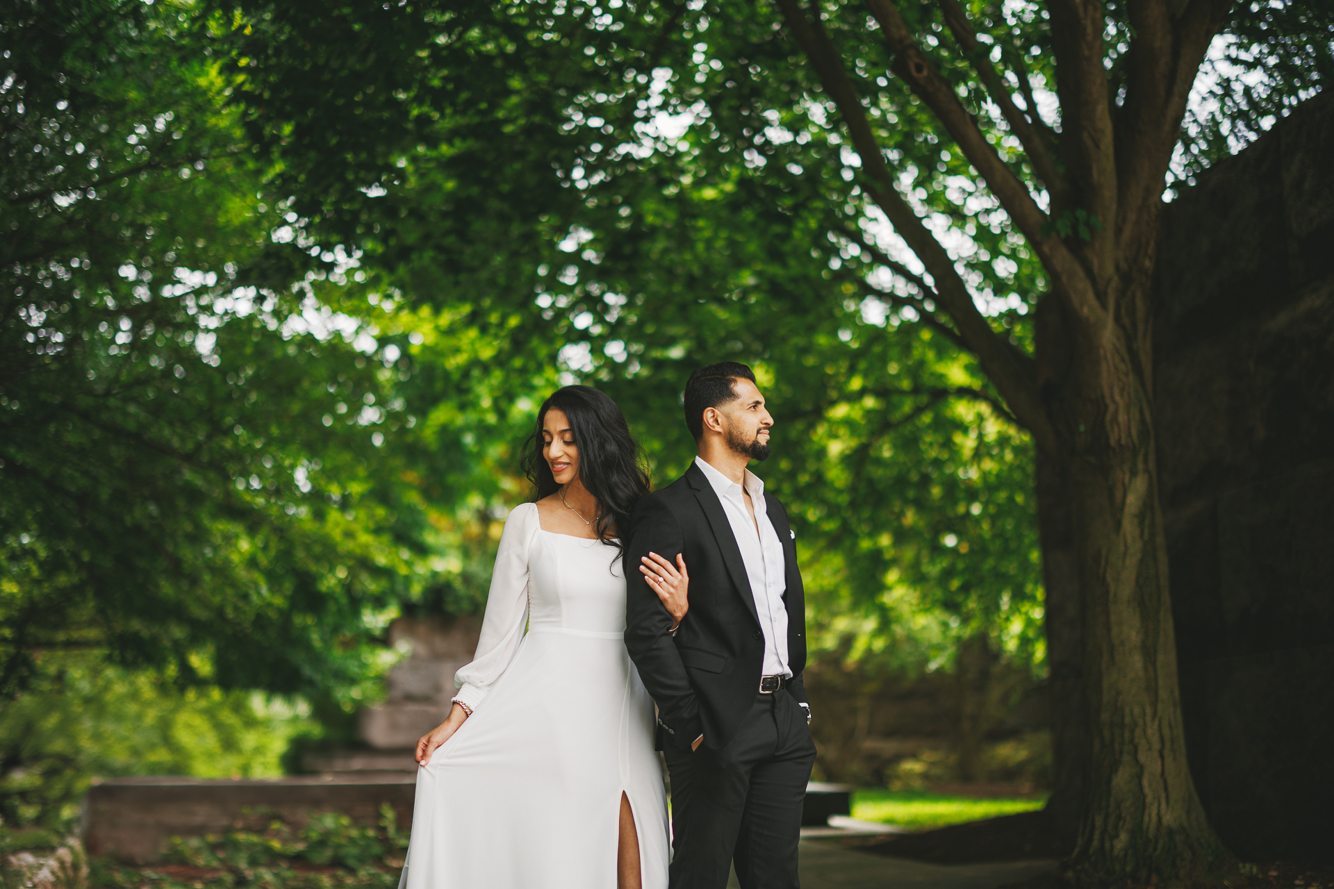 A woman in a white dress and a man in a black suit stand next to each other outdoors under a tree, as if part of an auto draft scene. The woman delicately holds part of her dress, while the man casually has his hands in his pockets.