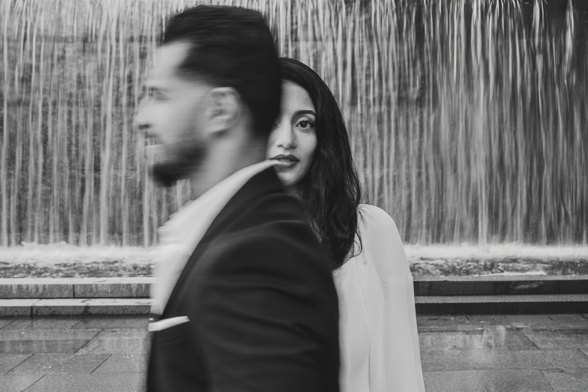 A woman stands in front of a waterfall, partially obscured by a man walking by. The image, reminiscent of an auto draft effect, is in black and white.