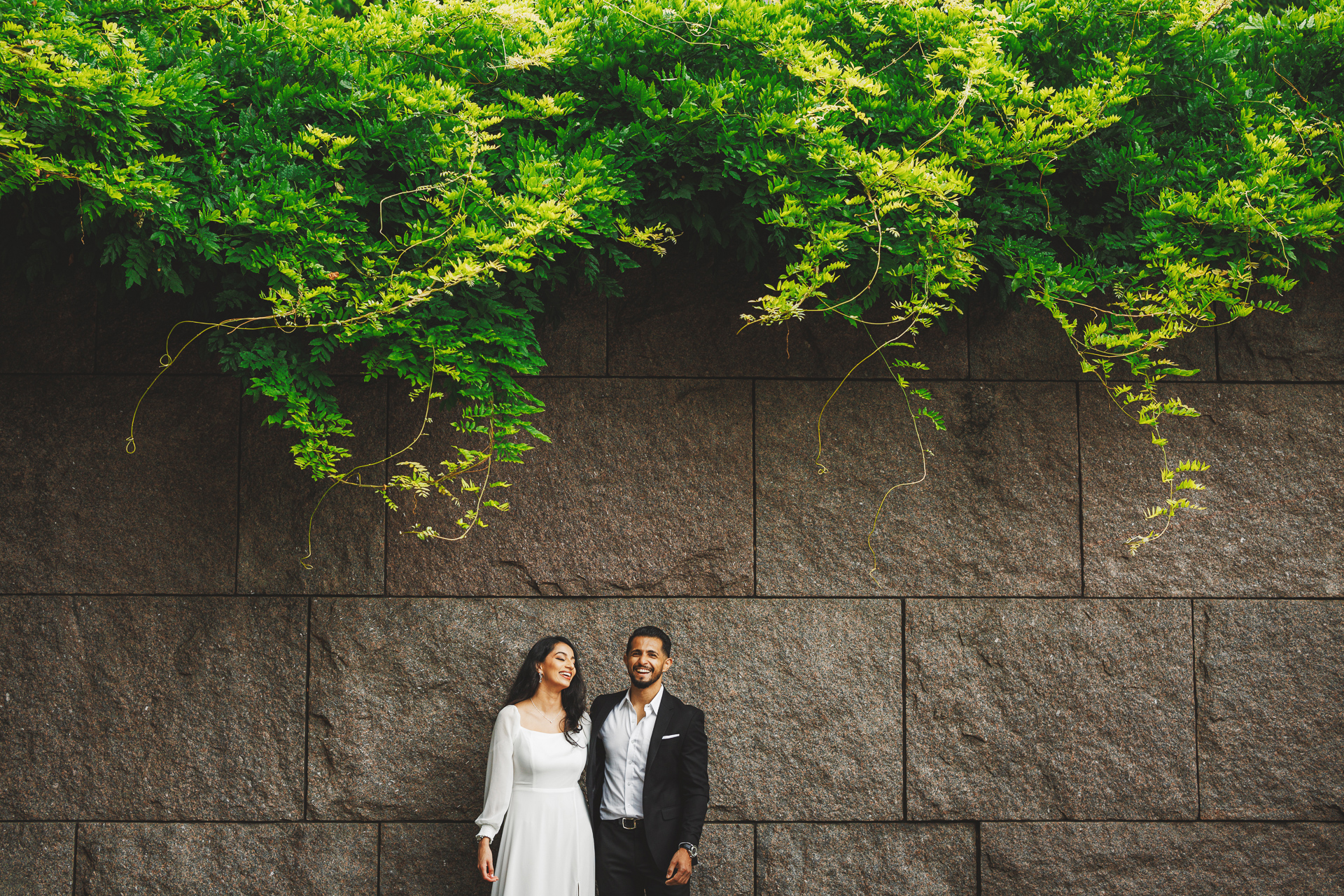 A man in a suit and a woman in a white dress stand side by side against a stone wall, lush green vines hanging above them, as if drafted into the perfect moment.
