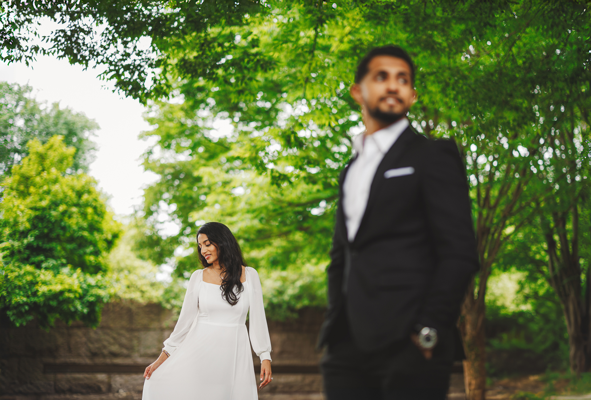 A man in a black suit stands in the foreground, while a woman in a white dress stands slightly behind him in the lush garden setting, seamlessly incorporating an auto draft of elegance.