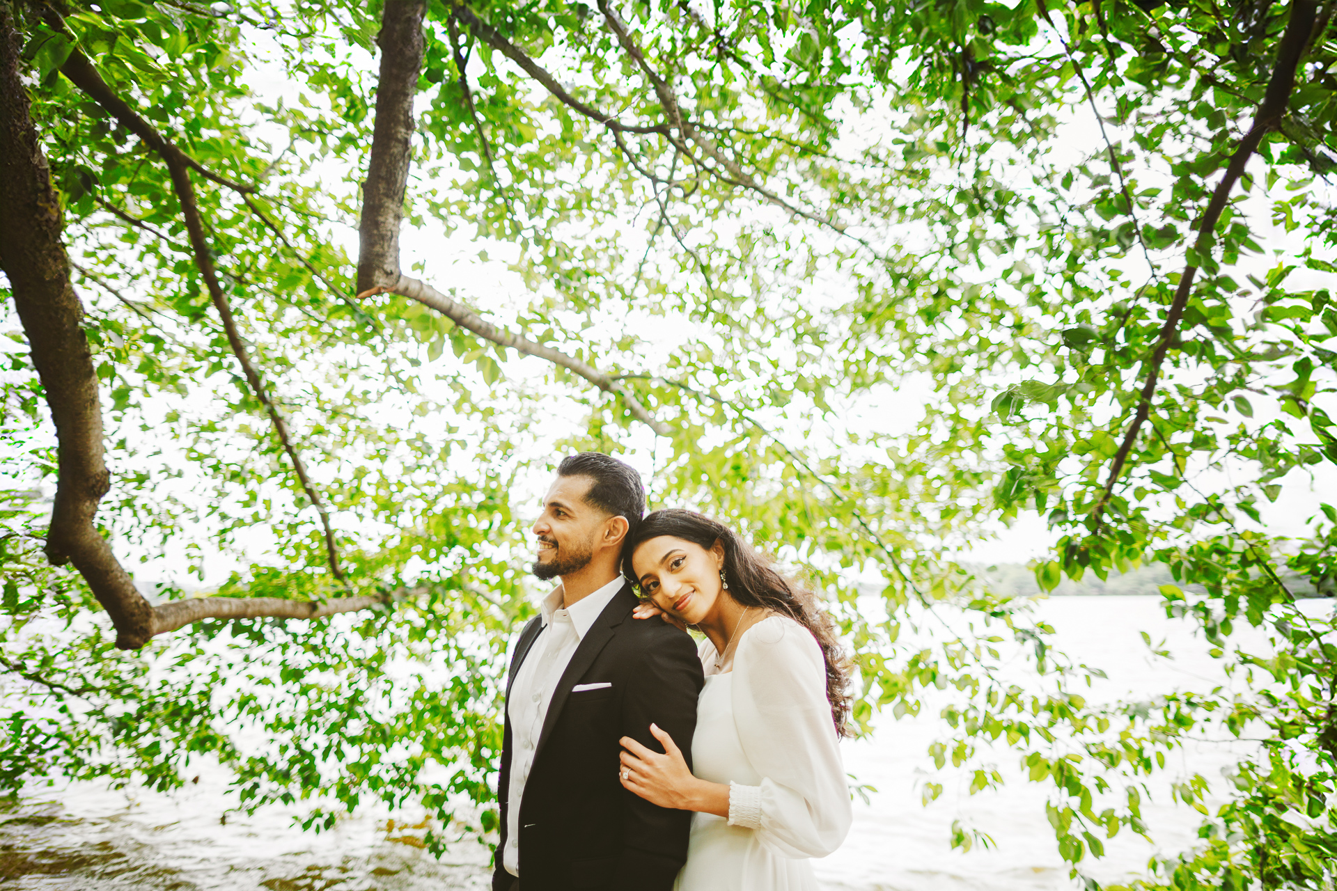 A woman in a white dress rests her head on a man in a black suit while standing under a tree with green leaves, as if posing for an Auto Draft moment.