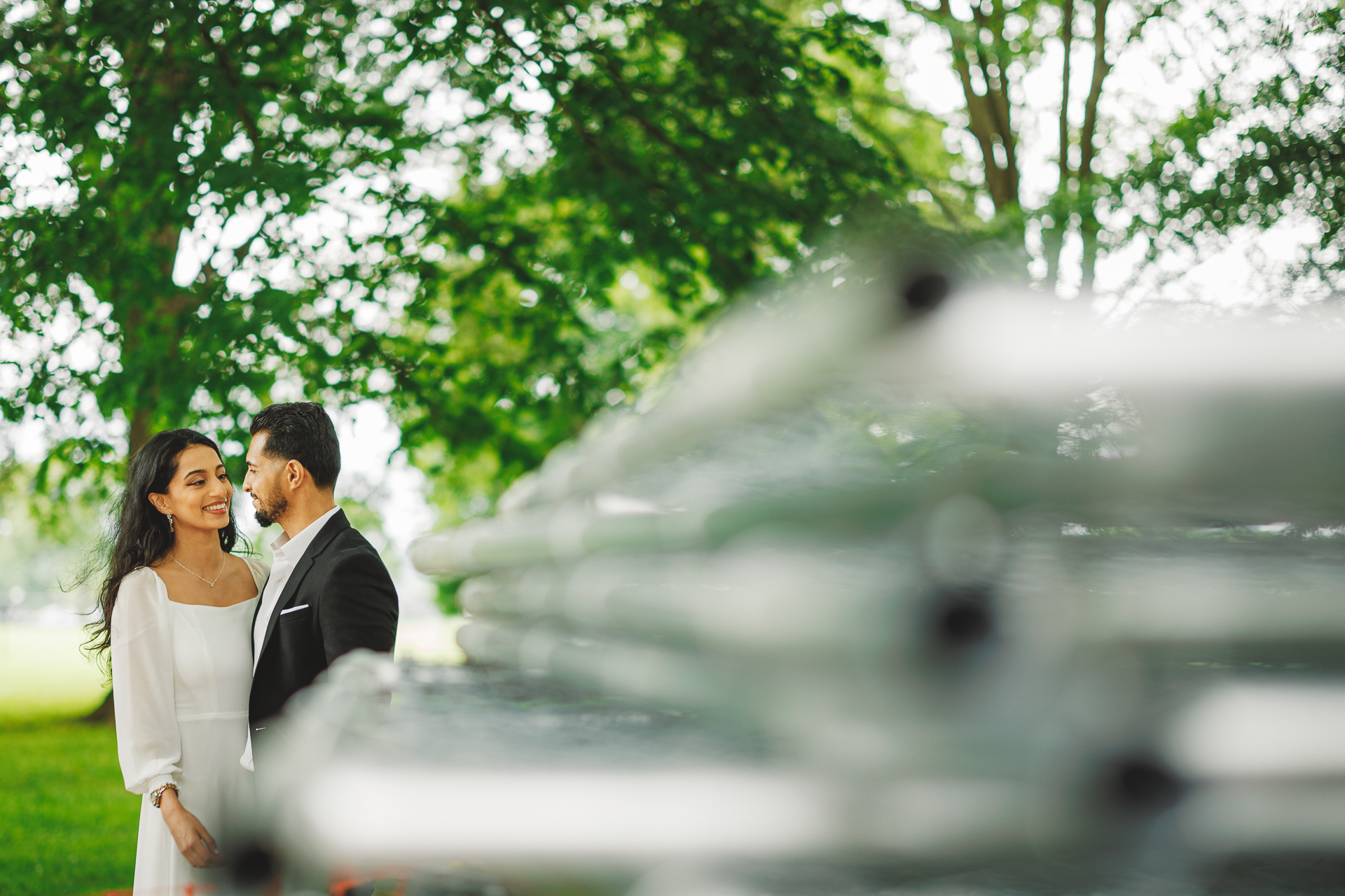 A couple is standing close together, smiling at each other with trees in the background. The image, resembling an auto draft of a perfect moment, is partially obstructed by a blurred fence in the foreground.
