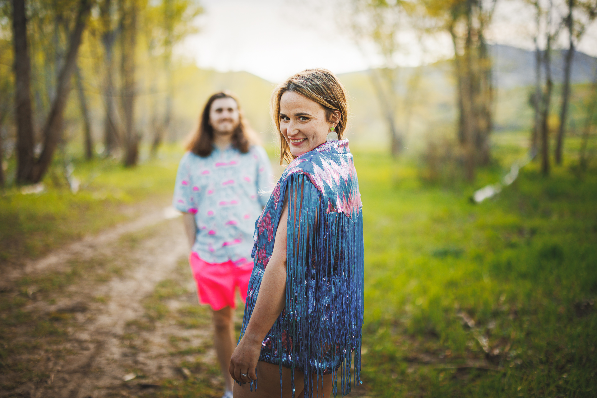 A woman wearing a fringed top smiles while standing on a forest path, auto draft in the gentle breeze. In the background, a man in colorful clothing adds to the vibrant scene.