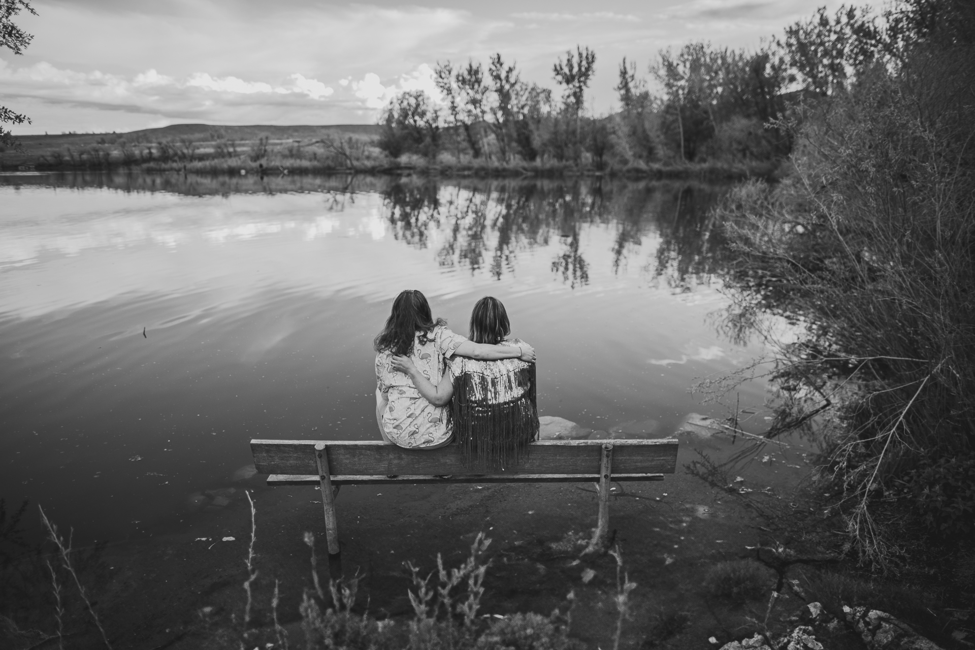 Two people sit closely on a bench near a calm lake, surrounded by trees and natural scenery. They are looking at the water, as if lost in an auto draft of memories. The image is in black and white.