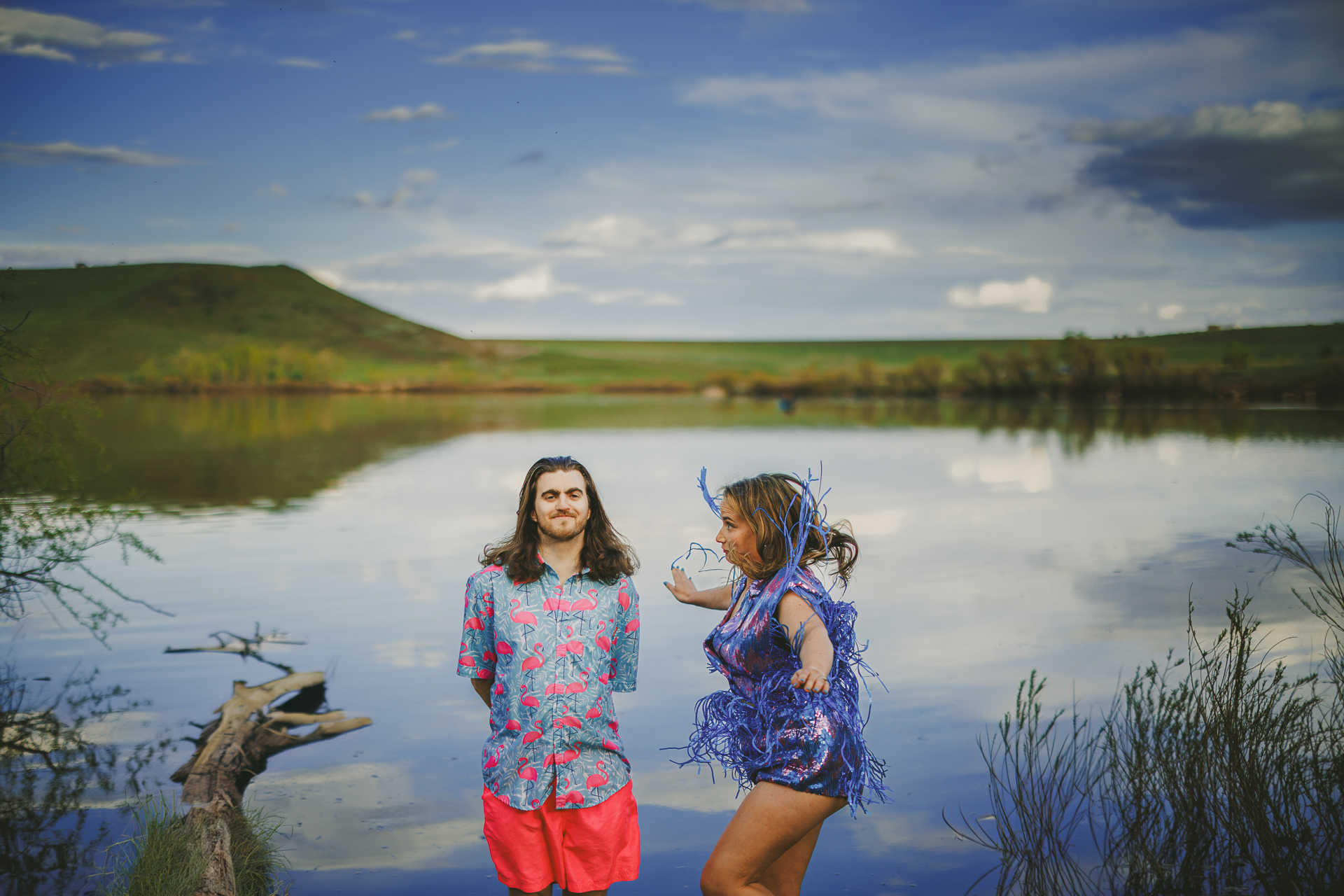 A man and a woman stand near a tranquil lake. The man wears a floral shirt and bright shorts, while the woman is in a shiny, festive outfit. Hills and a cloudy sky are visible in the background, creating an idyllic scene that seems almost like an auto draft of paradise.