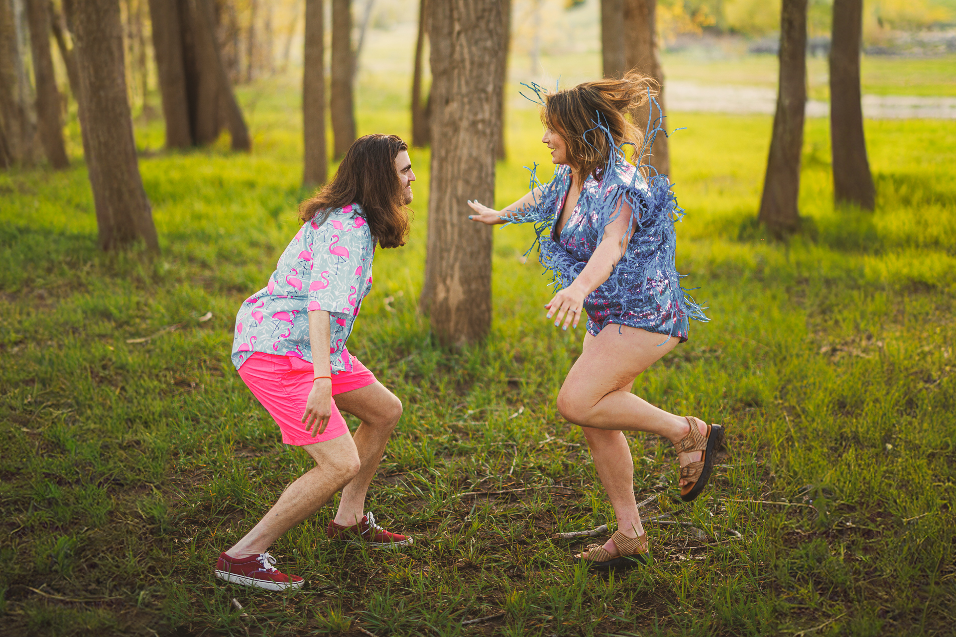Two people in brightly colored shirts and shorts playfully interact in a wooded area with tall trees and green grass. One person lunges toward the other, who is mid-jump with arms extended, like a perfectly executed auto draft of joy and spontaneity.