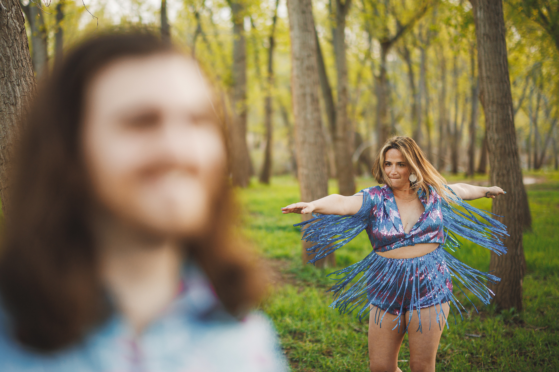 Person in foreground with long hair and blurred face. In the background, a person in a colorful fringed outfit stands with arms outstretched among the trees, perfectly capturing the auto draft of nature's beauty.