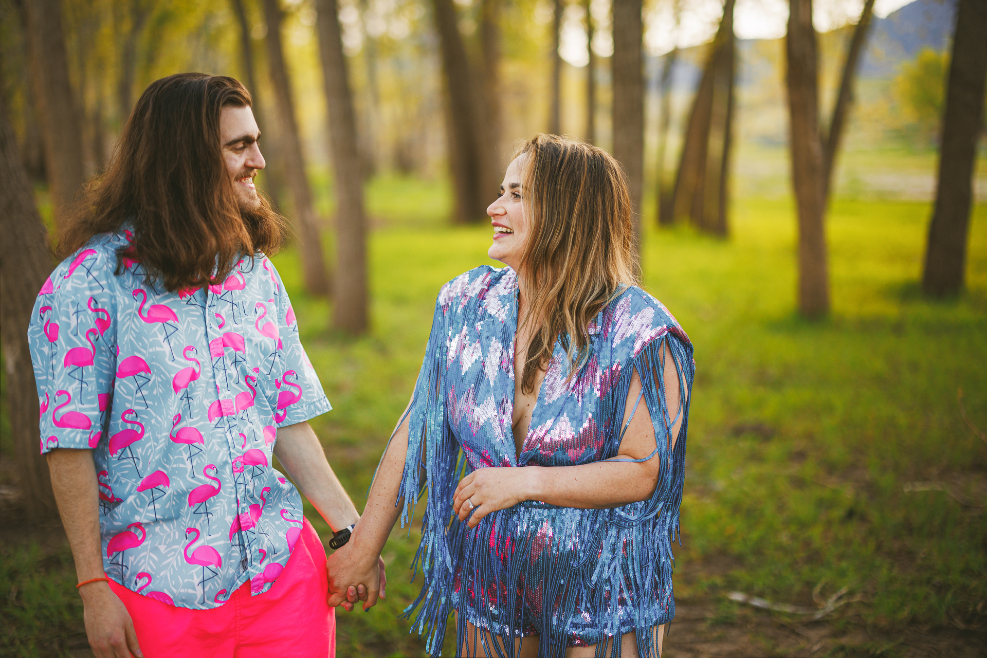 A man and a woman smiling at each other, holding hands in a forested area. The man, wearing a flamingo-patterned shirt, seems effortlessly stylish while the woman complements him beautifully in her blue fringed outfit. Their connection feels as spontaneous and perfect as an auto draft of happiness.