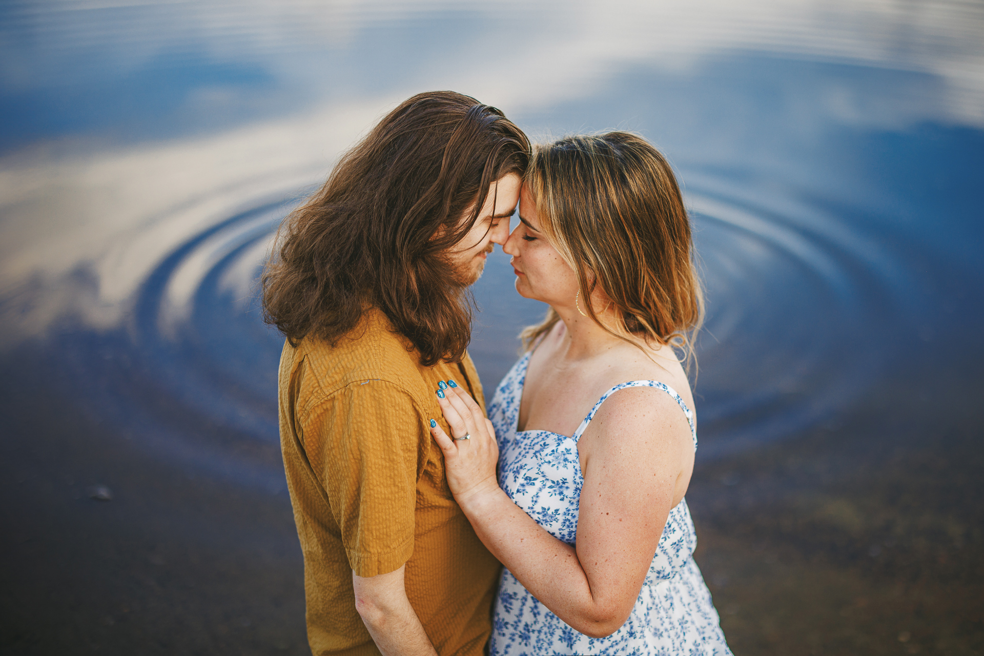 Two individuals stand close to each other near a calm body of water, creating gentle ripples, as they gaze into each other's eyes. The person on the left has long hair and wears a mustard-colored shirt while the other wears a light blue patterned dress, their connection seeming almost auto draft in its effortless perfection.