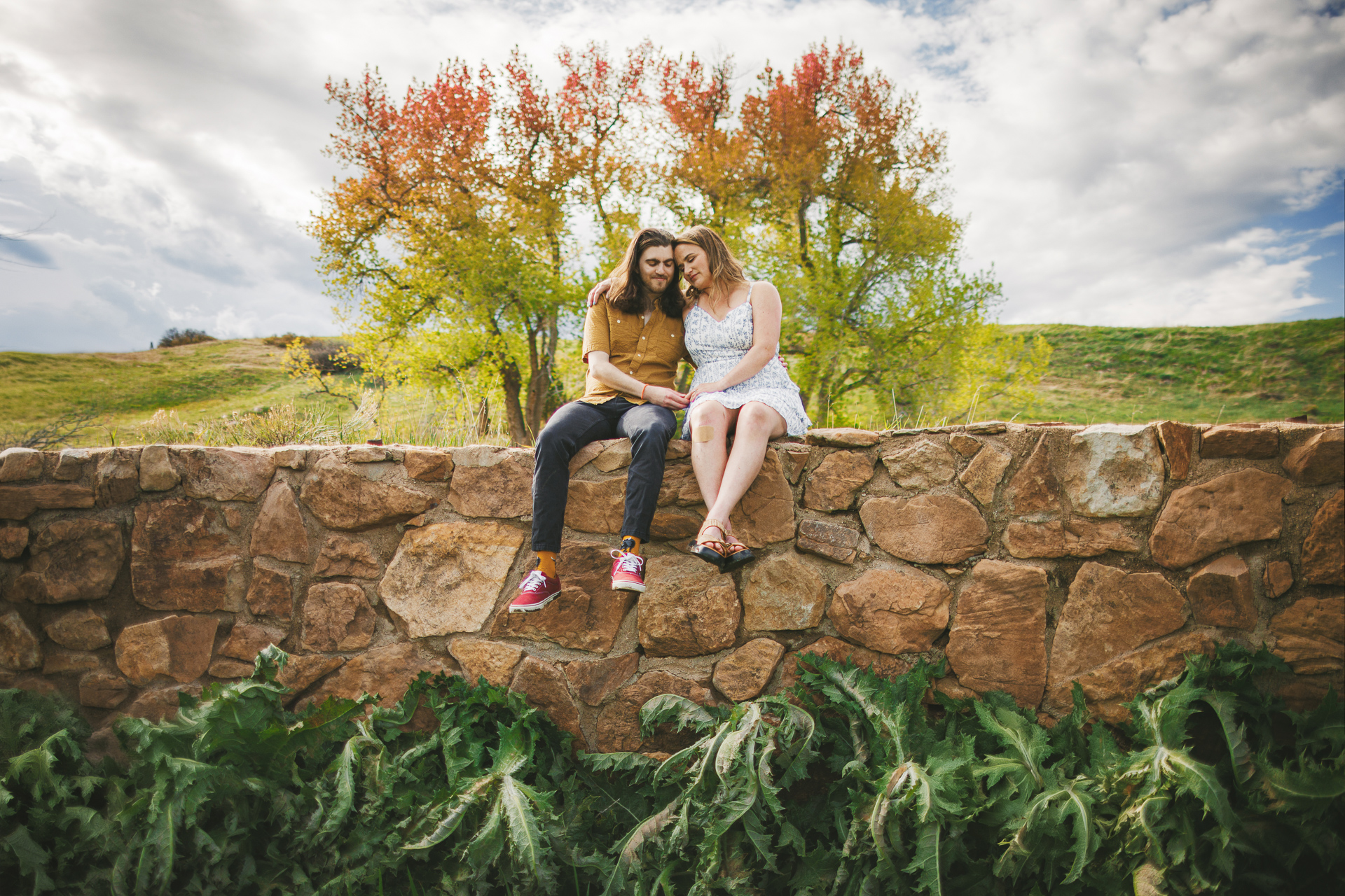 Two people sit together on a stone wall with green plants below and trees with autumn foliage in the background, their quiet moment framed by the partly cloudy sky—an effortless blend like an auto draft of nature's scenery.
