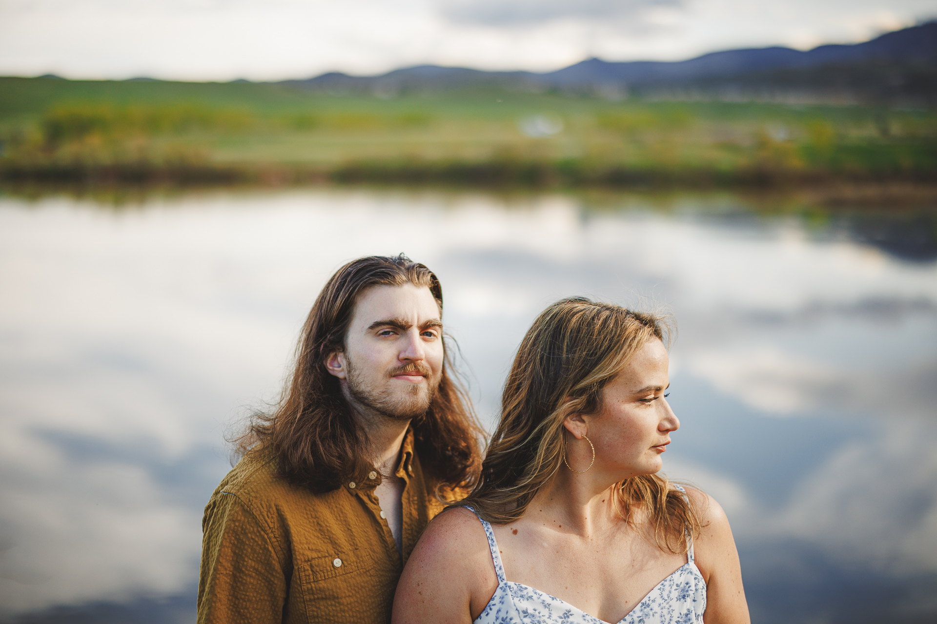 Two individuals stand in front of a serene lake with a blurred backdrop of mountains and sky. The person on the left, looking like an auto draft from a nostalgic memory, has long hair and wears a button-up shirt; the person on the right has blonde hair and wears a patterned dress.