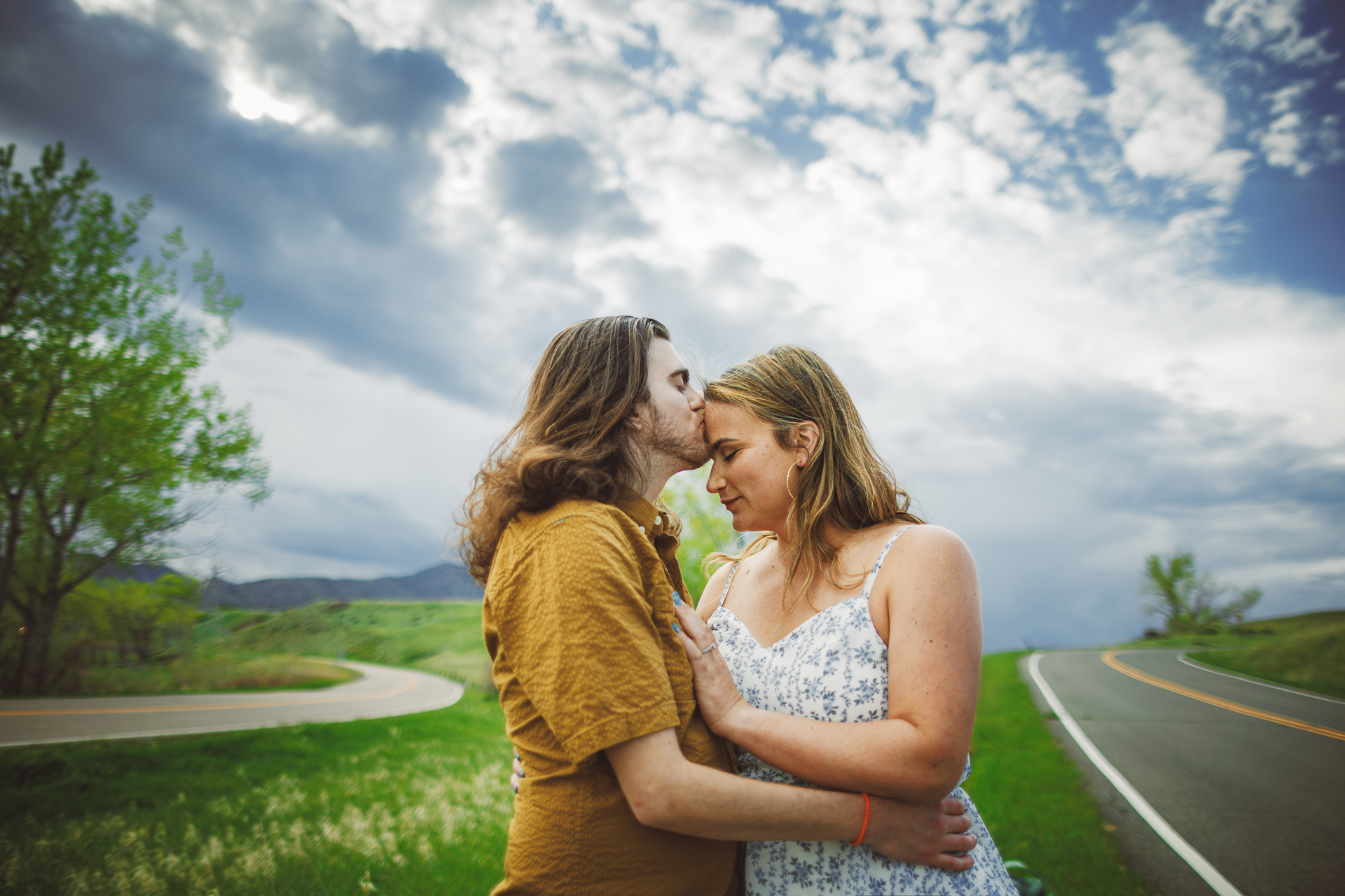 A couple stands on a curved road surrounded by greenery and trees, sharing a tender moment under a partly cloudy sky. The man in a mustard shirt and the woman in a floral dress look as if they were plucked from an Auto Draft of life's beautiful moments.