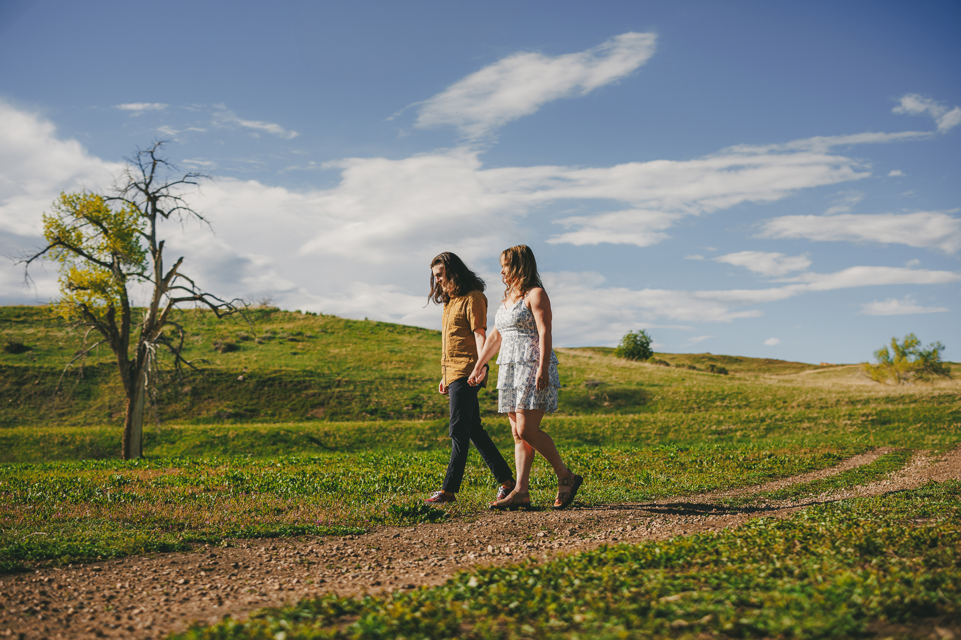 Two people walk hand-in-hand along a dirt path in a grassy field under a blue sky with scattered clouds, as if scripted by an auto draft of nature's perfection.